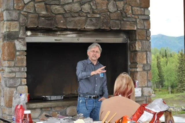 Man speaking at an outdoor gathering by a stone fireplace with people seated, surrounded by trees and mountains in the background.