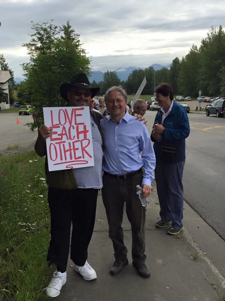 Two men standing on a sidewalk, one holding a sign that says 'LOVE EACH OTHER', with other people and parked cars in the background.