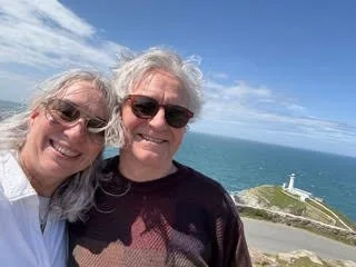 A smiling older couple taking a selfie near the coast with a lighthouse on a rocky shoreline in the background.