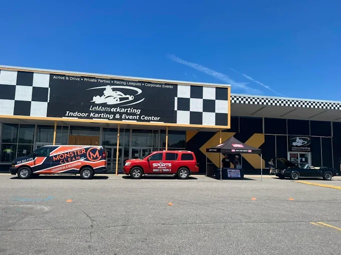 Front view of LeMans karting Indoor Karting & Event Center with parked vehicles, including a Monster Cleaner van, a red SUV, a black car, and a tent outside on a sunny day.