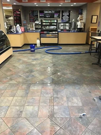 Inside a coffee shop, with colorful tiled floor, a blue vacuum hose lying on the floor, and a counter with menu boards and equipment in the background.