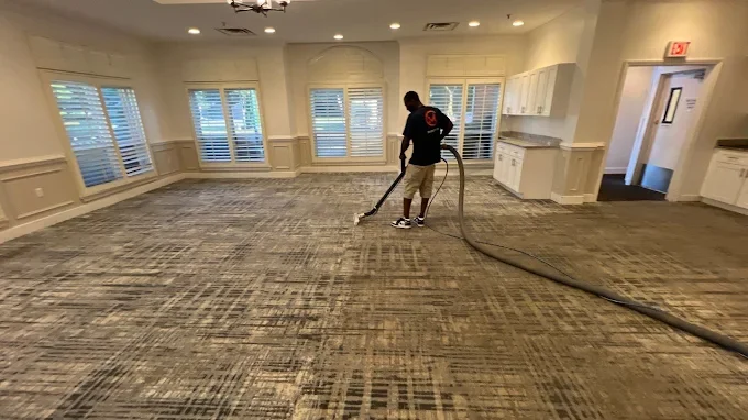 A man vacuuming a large, empty room with multiple windows and white cabinets.