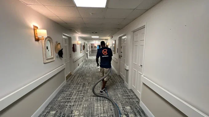 A man cleaning a hallway with a hose in what appears to be a hotel or office building corridor.