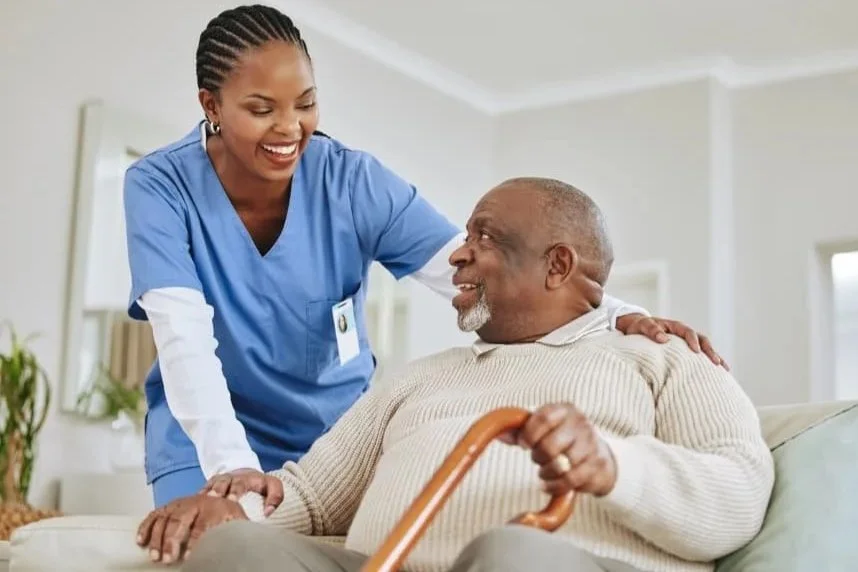 A nurse smiling and talking with an elderly man sitting on a sofa, holding a cane in a bright room.