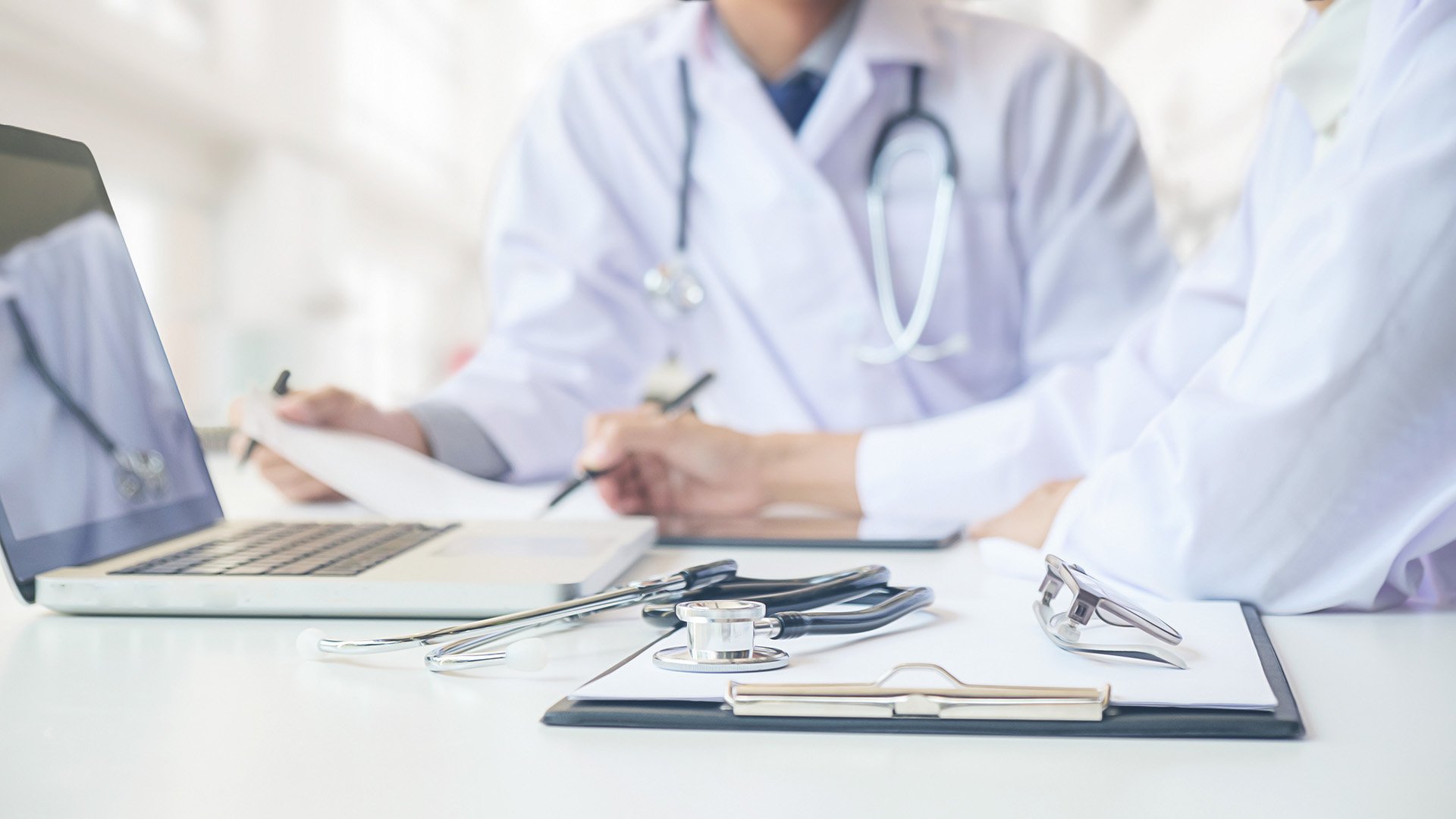 Medical professionals in white coats with stethoscopes at a desk with a laptop, clipboard, and medical tools.