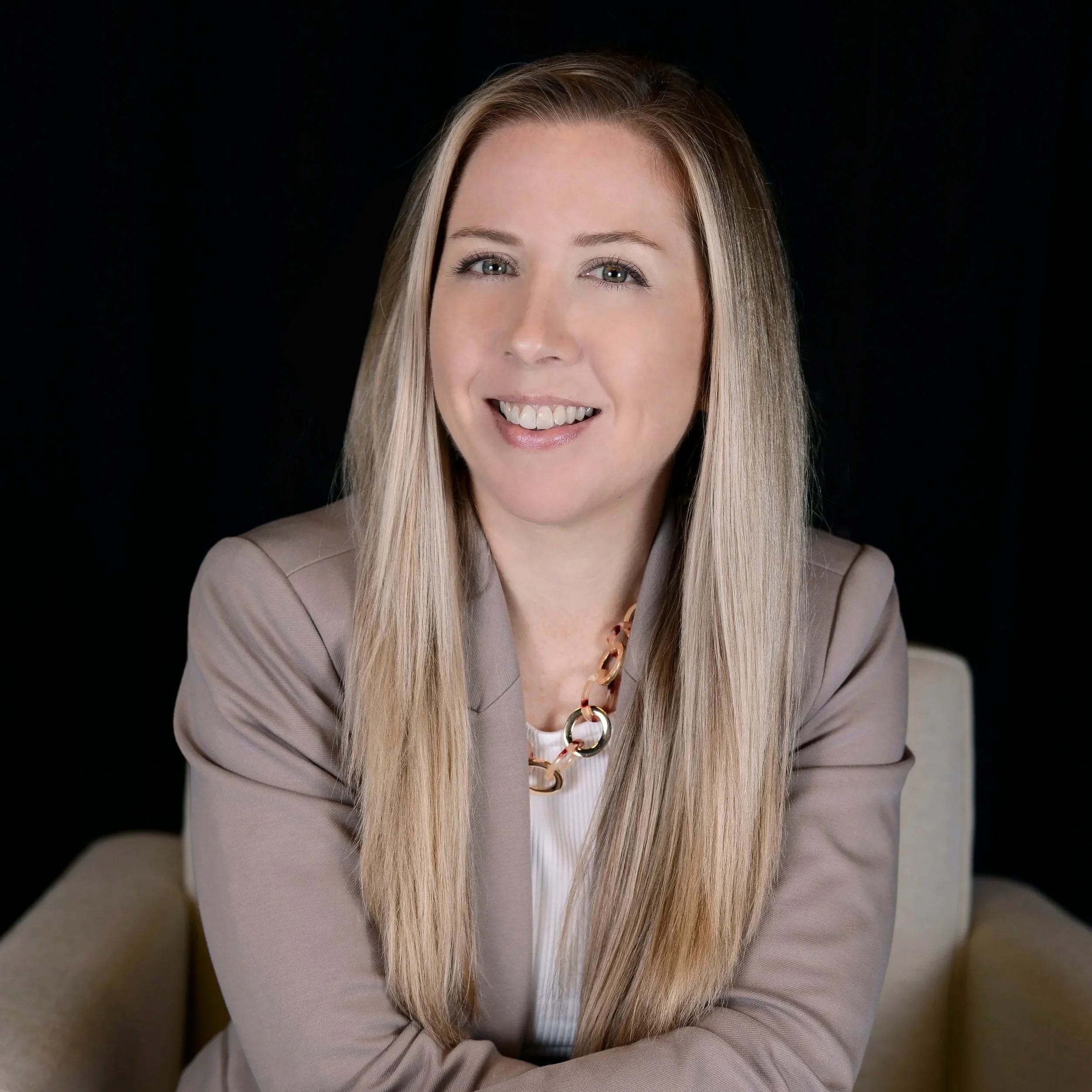 A woman with long blonde hair, wearing a beige blazer and a chunky chain necklace, smiling at the camera against a black background.