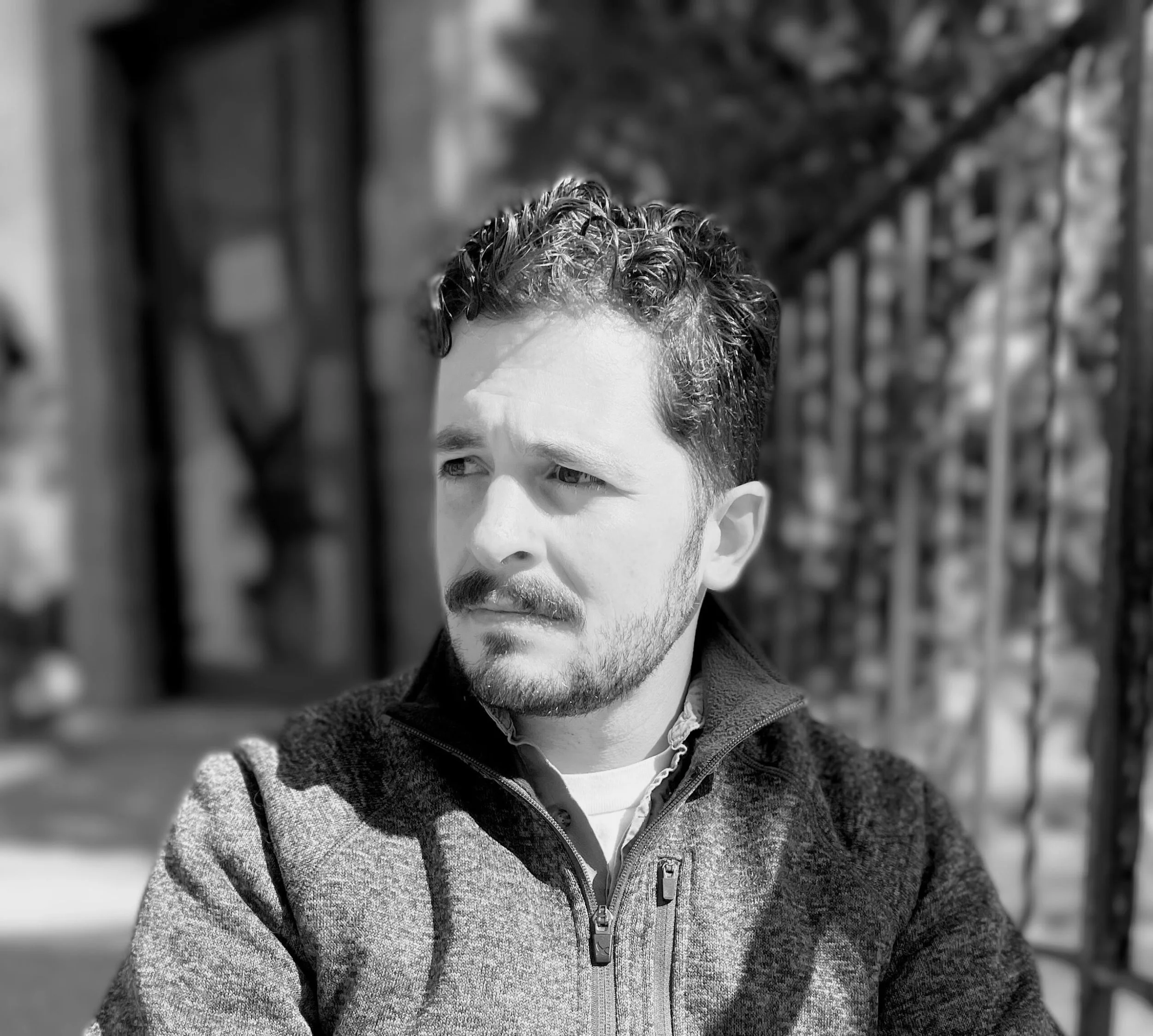 Black and white photo of a man with curly hair and a beard, looking to the side, sitting outside near a metal fence.