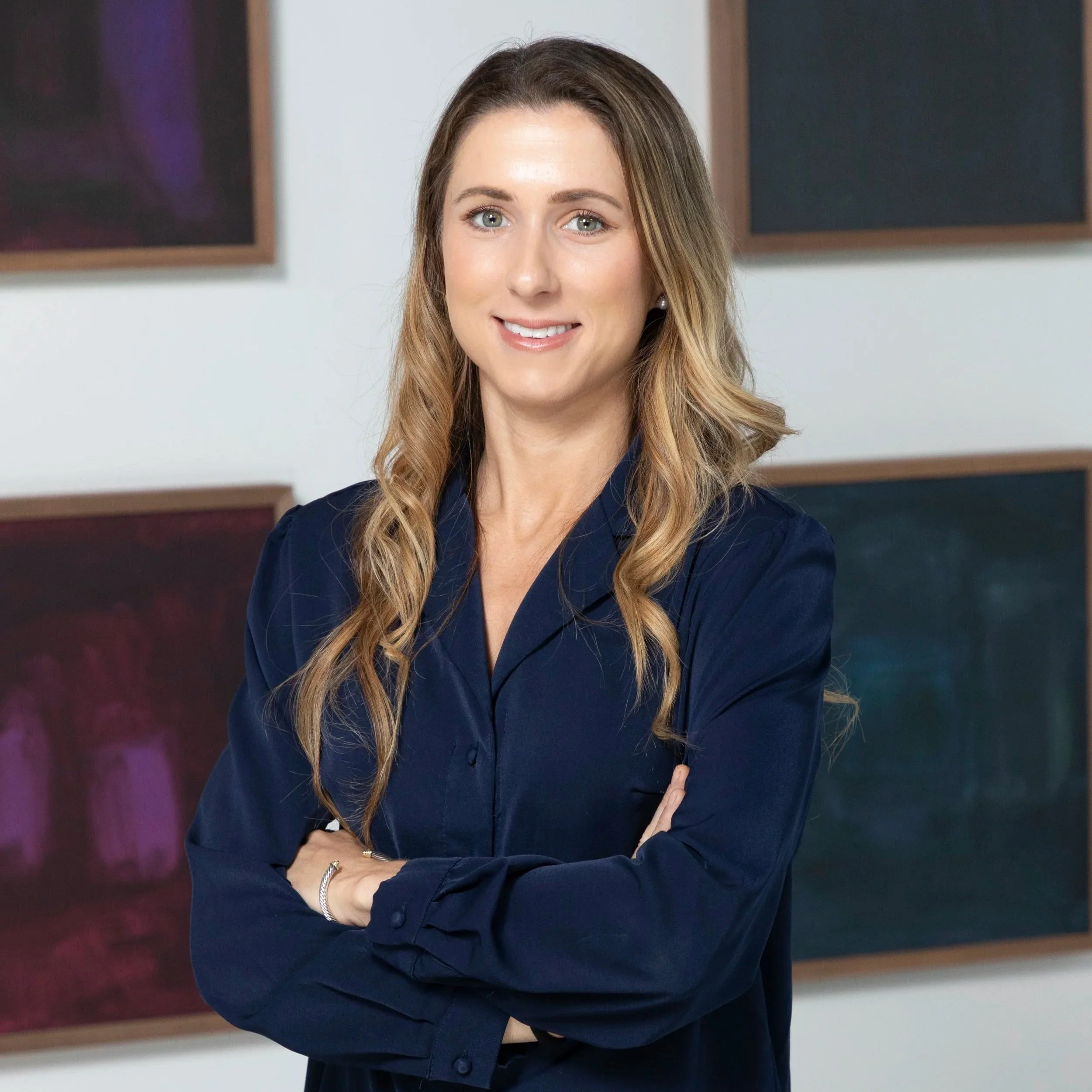 A woman with blonde, wavy hair, wearing a navy blue blouse, smiling with arms crossed, standing in front of abstract artwork.