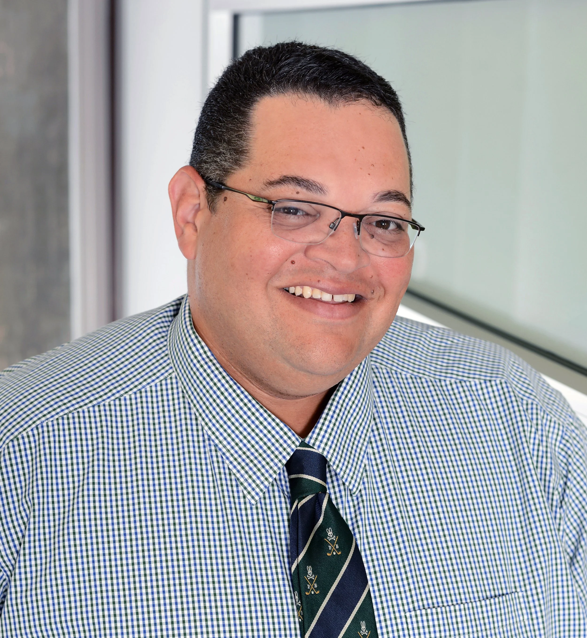 A smiling man with glasses wearing a checkered shirt and a striped tie, sitting in front of a window.