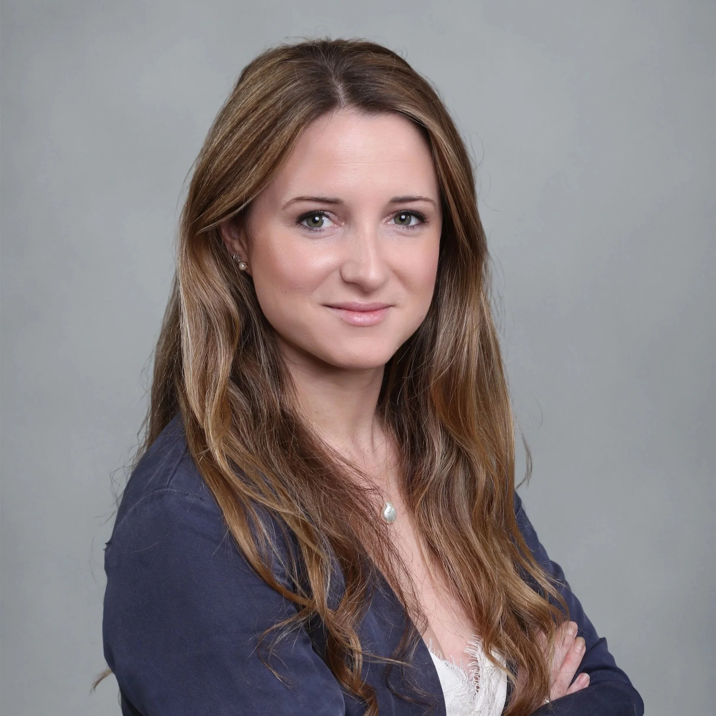 A woman with long, wavy brown hair and wearing a navy blazer, posing with her arms crossed and smiling gently against a plain gray background.