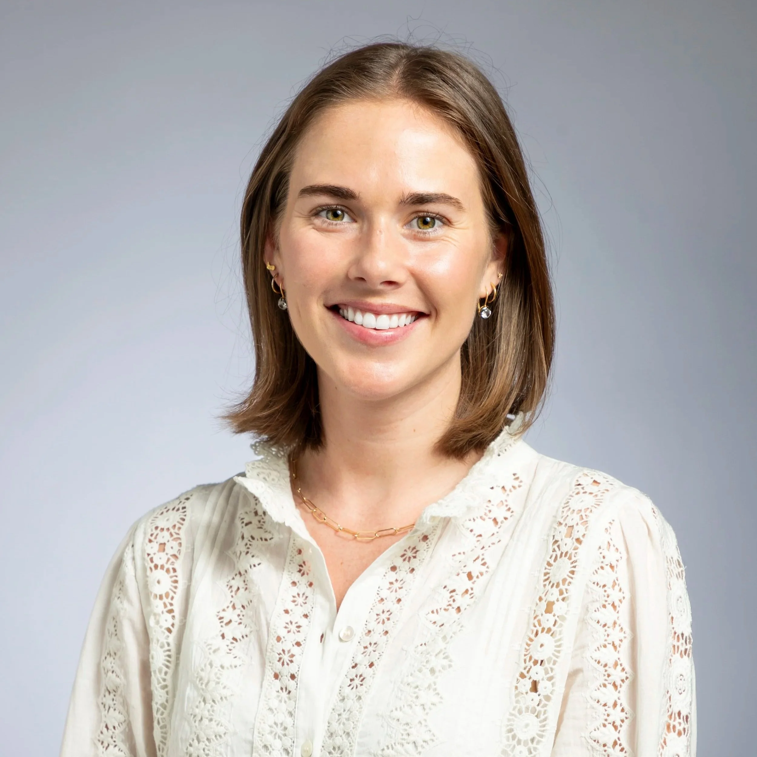 A woman with shoulder-length brown hair, wearing a white lace blouse and gold jewelry, smiling at the camera against a black background.
