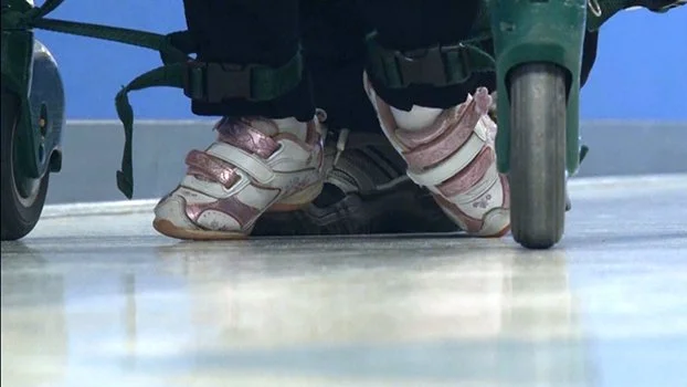 Child in pink and white sneakers sitting in a wheelchair with wheels visible, on a light-colored floor.
