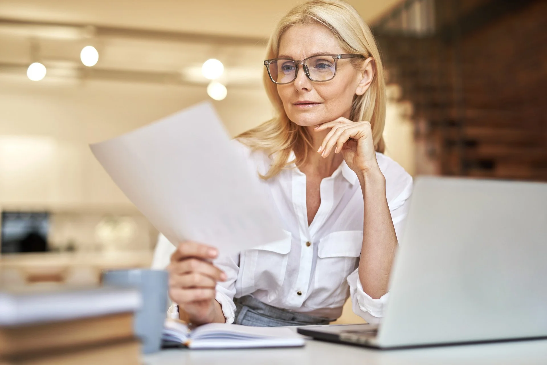 A blonde woman wearing glasses and a white shirt, sitting at a desk, reading a piece of paper, with a laptop and some books in front of her, in an office or study environment.