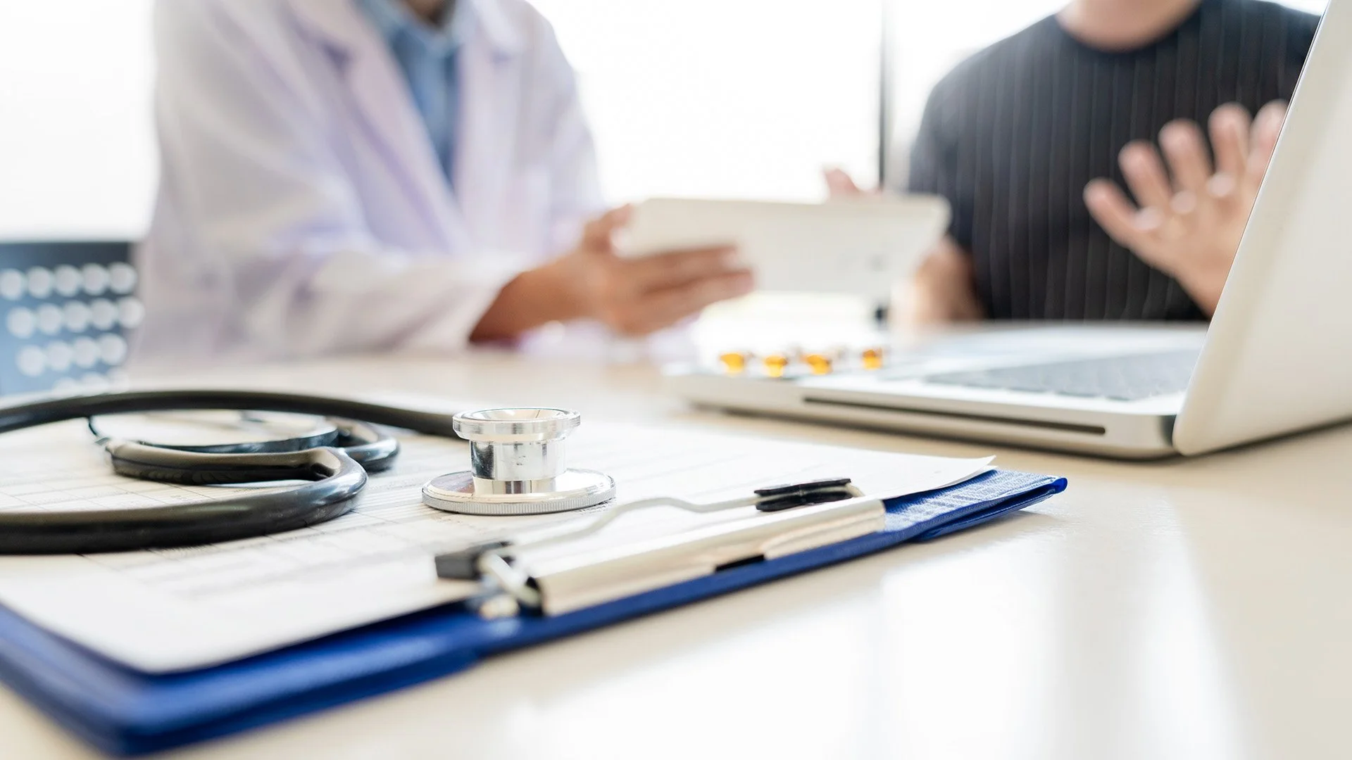 Close-up of a stethoscope, clipboard, and medical records on a table with a doctor and patient in the background, blurred.