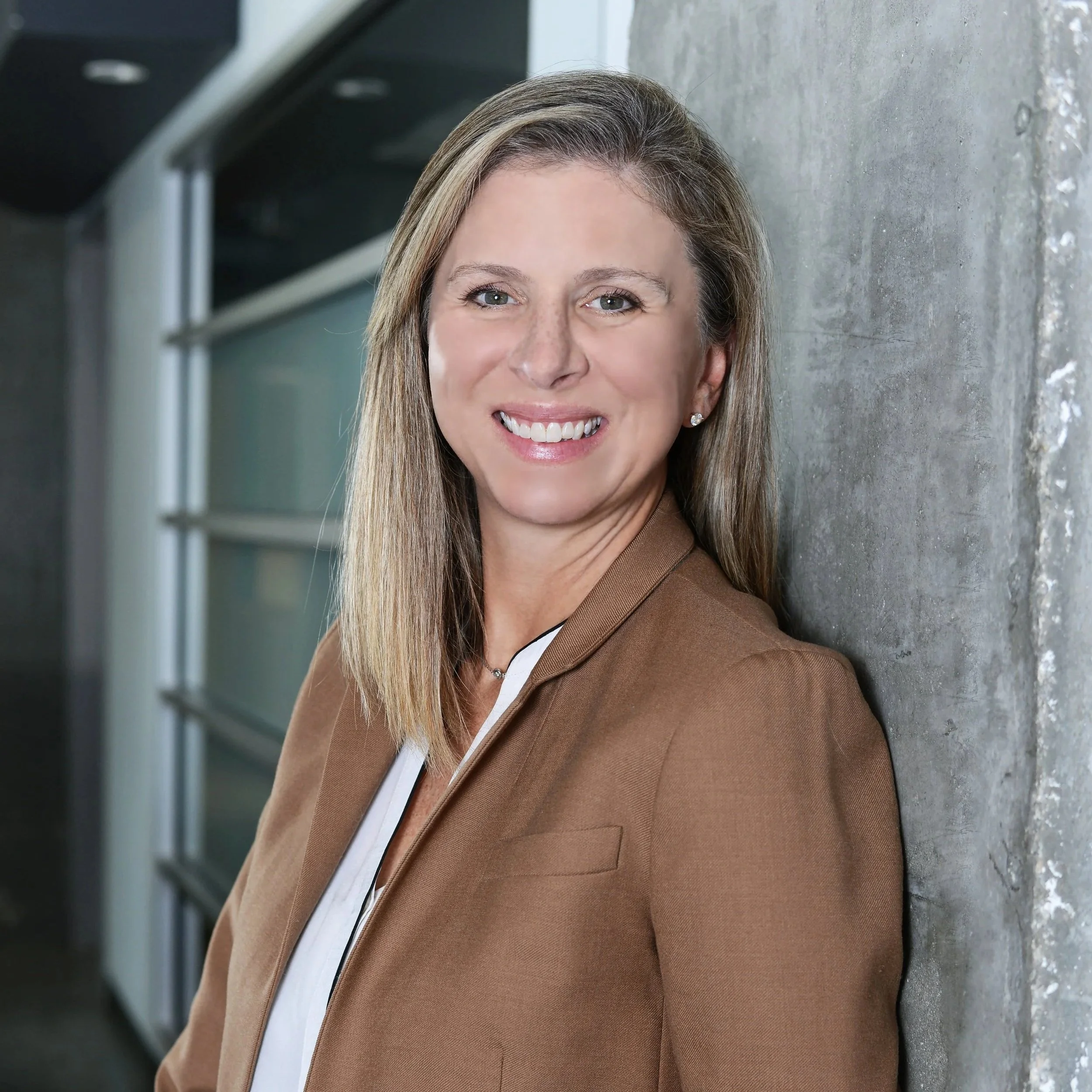A smiling woman with shoulder-length blonde hair, wearing a brown blazer and white top, leaning against a concrete wall in an office setting.