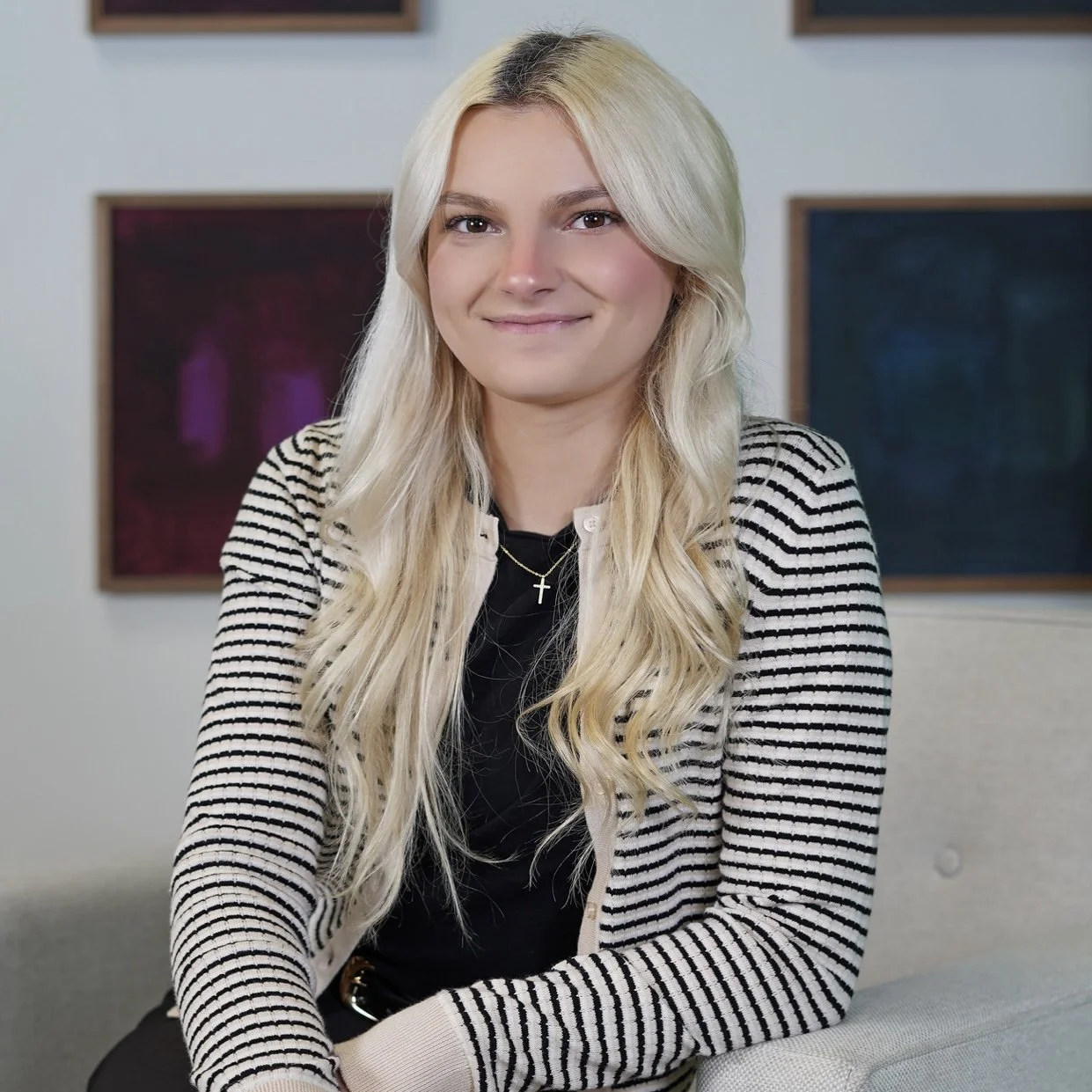 A young woman with long blonde hair, wearing a striped blazer, black top, and a cross necklace, sitting and smiling in an indoor setting with framed artwork on the wall behind her.