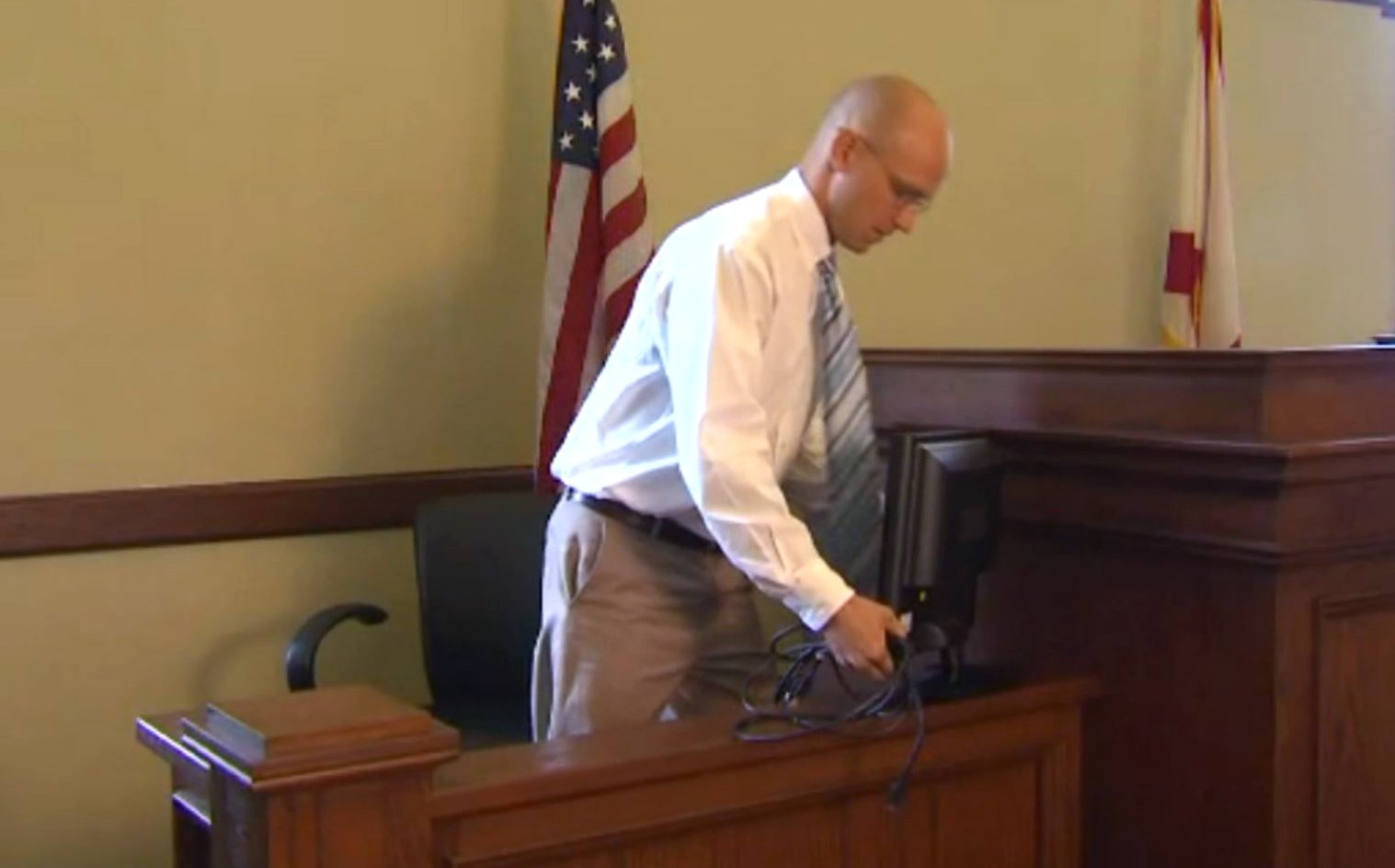 A man in a white shirt and tie working with computer cables in a courtroom, with an American flag behind him.