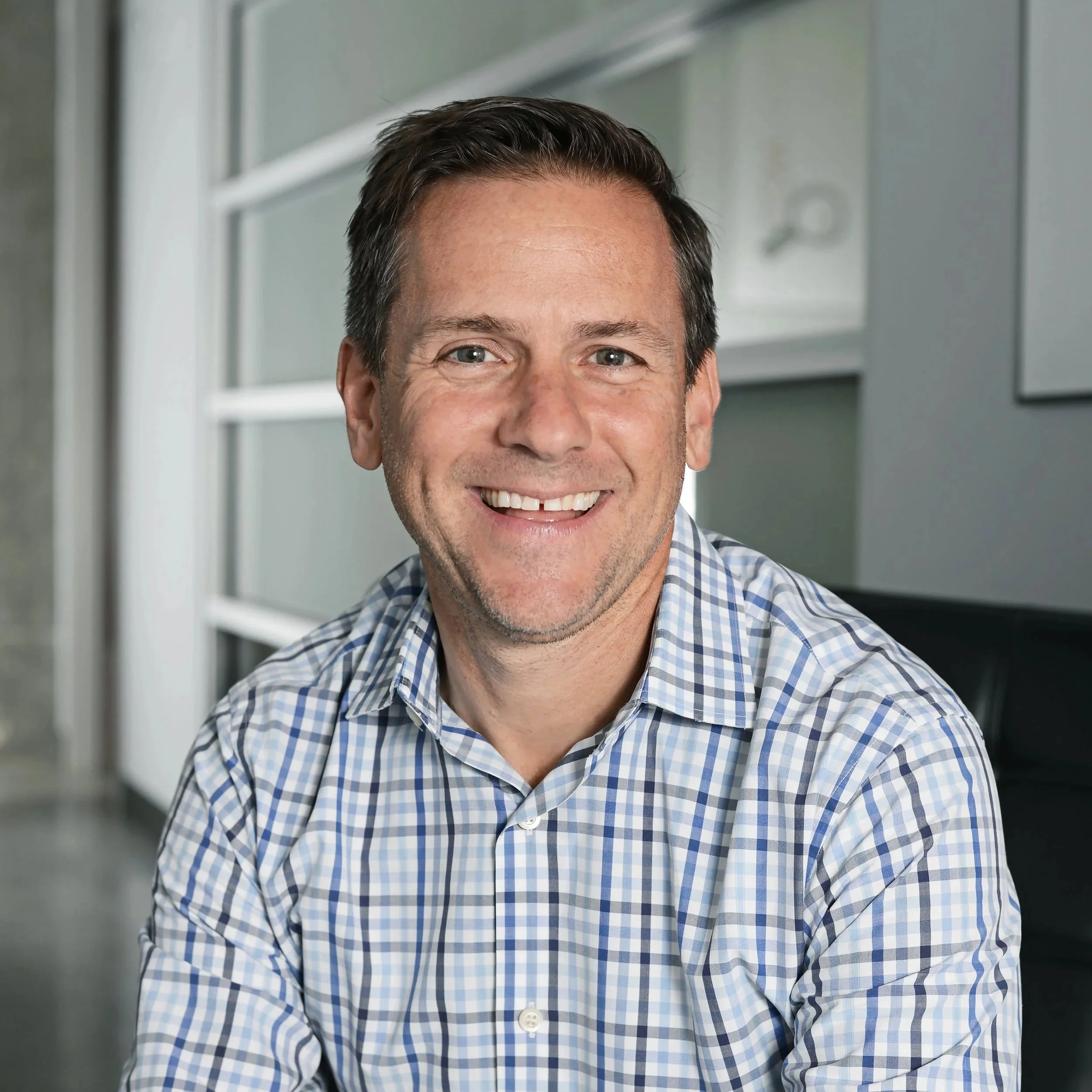 A smiling man with short dark hair wearing a blue and white checkered shirt, seated indoors with a blurred background.