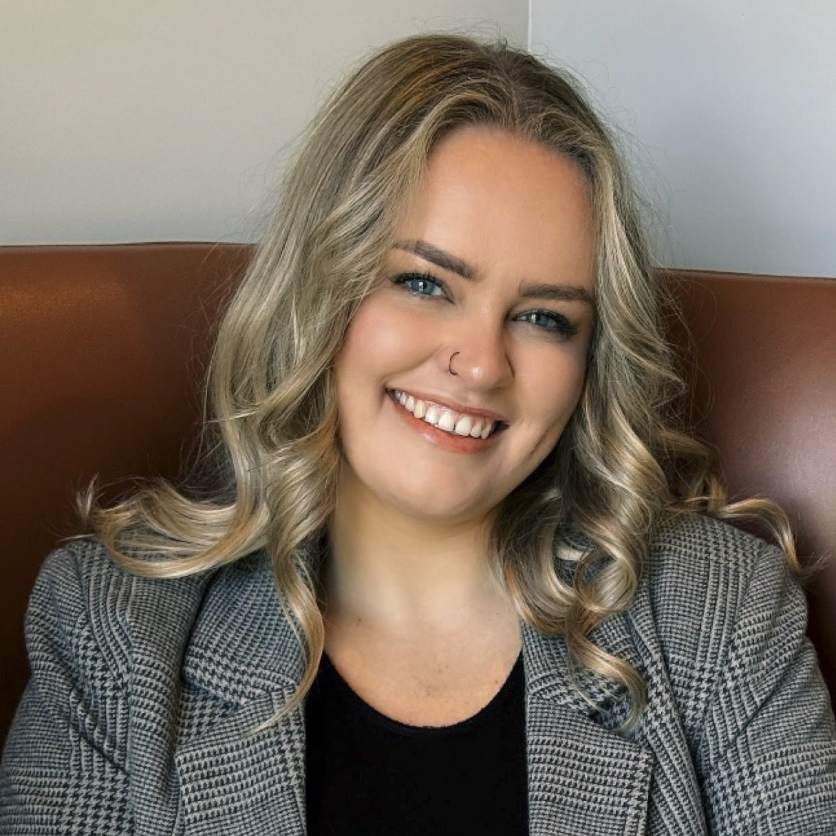 A smiling woman with blonde curly hair, blue eyes, and a nose piercing, wearing a gray jacket and black top, sitting on a leather chair.