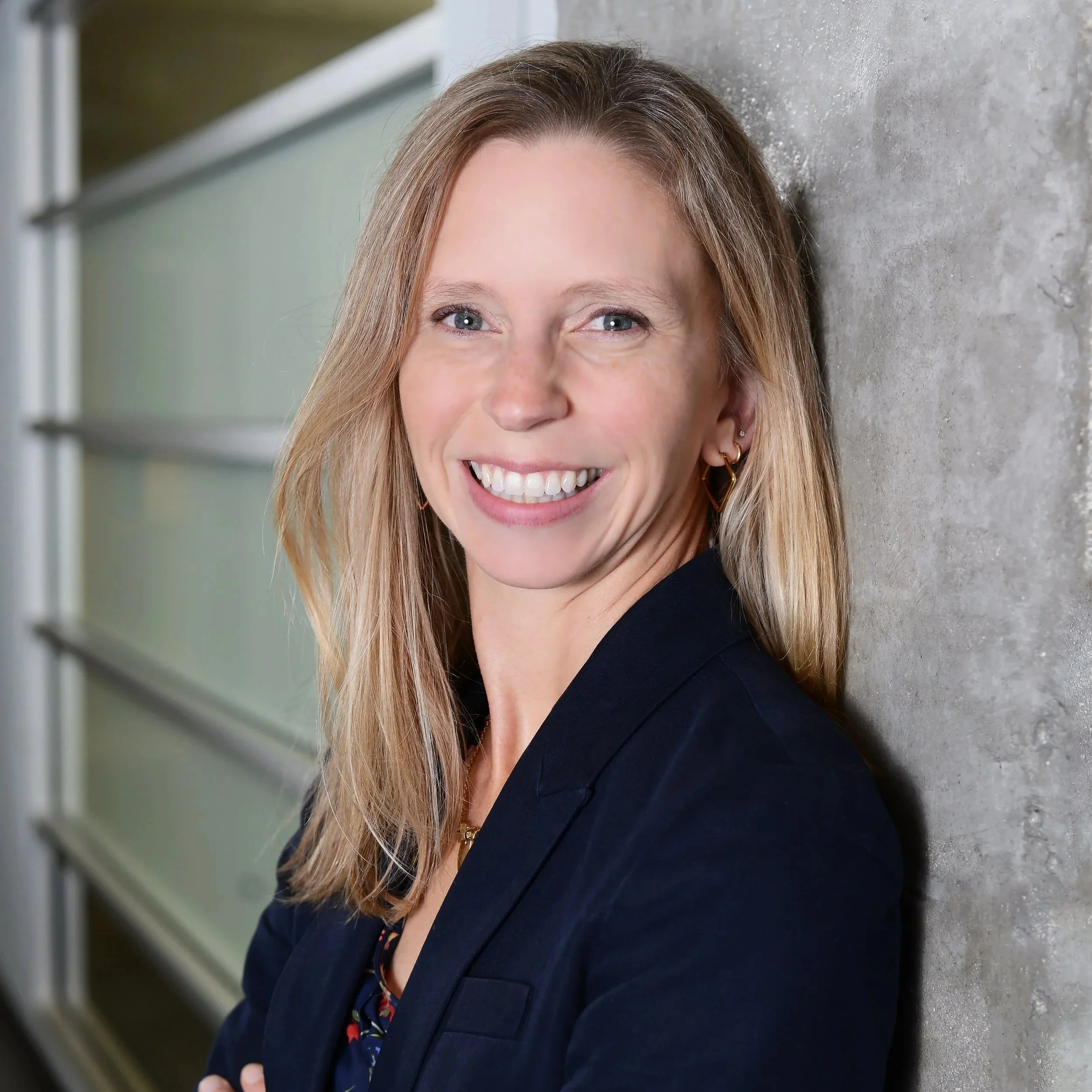 Portrait of a woman with blonde hair and blue eyes smiling, wearing a black blazer, gold earrings, and a patterned blouse, leaning against a concrete wall with metal shelving in the background.