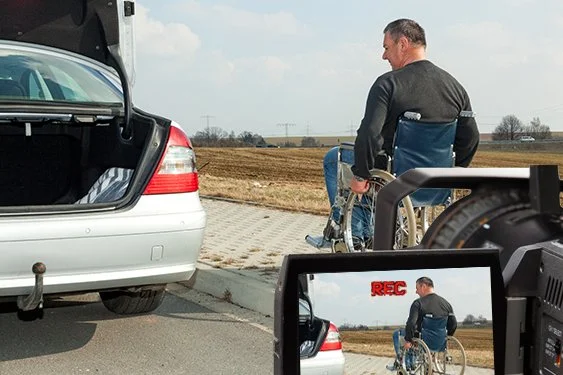 A man in a wheelchair is on the side of the road next to a silver car with its trunk open, while a camera records him from a monitor.