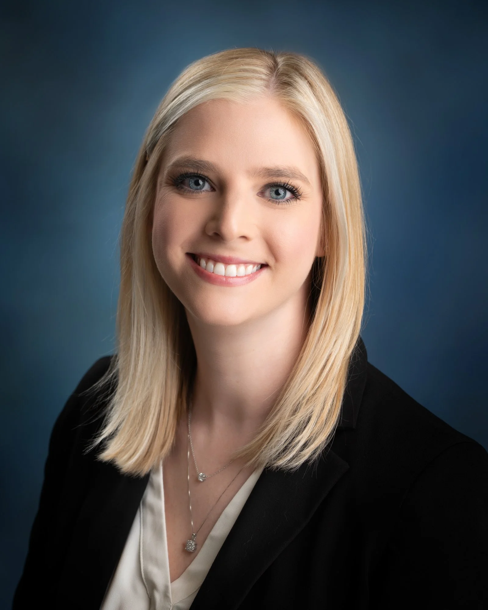 Headshot of a blonde woman with blue eyes smiling, wearing a black blazer, silver necklace, and a white blouse against a blue background.