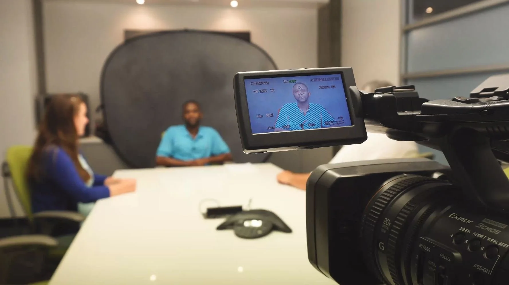 A camera capturing a video interview of two people in a conference room; a woman with long brown hair in a blue outfit and a man in a blue shirt sit at a table, with a backdrop behind them.