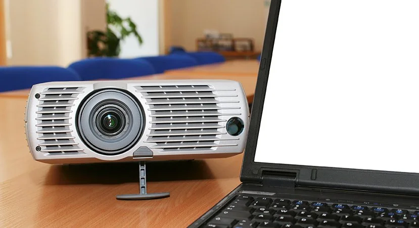 A white projector on a wooden table next to an open laptop with a blank white screen in a conference room.