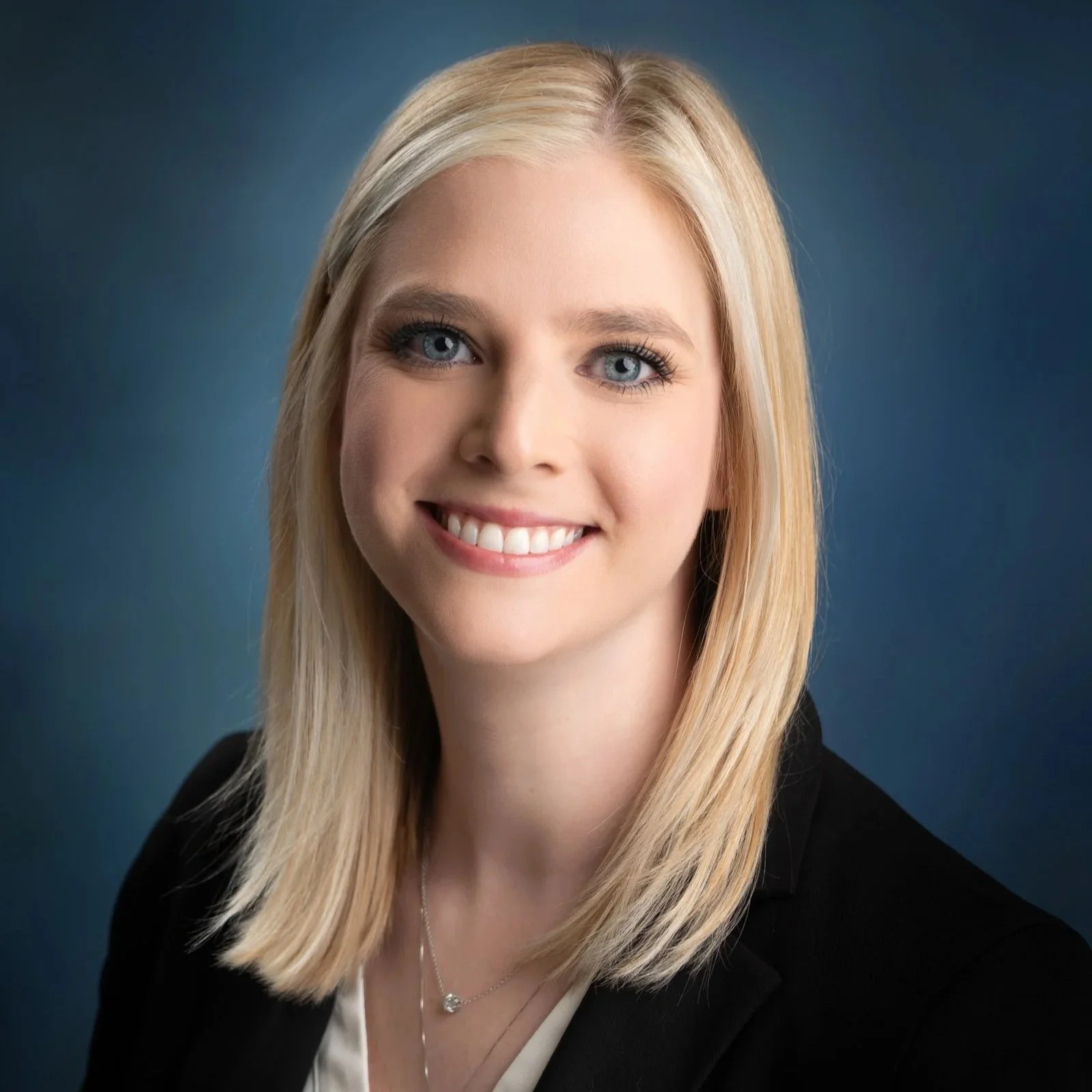 A professional headshot of a young woman with shoulder-length blonde hair, blue eyes, and a bright smile, wearing a black blazer and a delicate necklace, against a dark blue background.