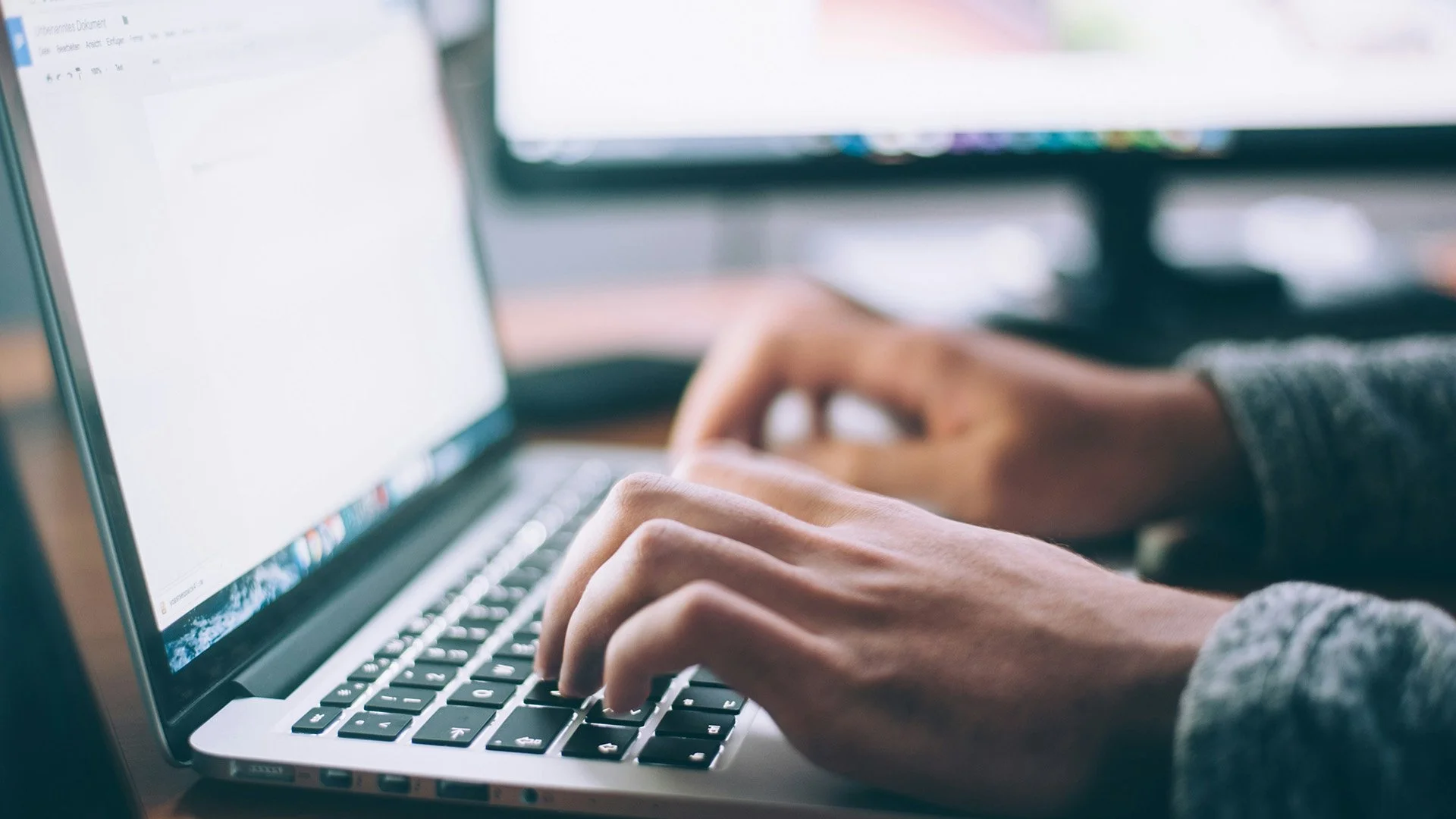 Close-up of a person's hands typing on a laptop keyboard with a blurred screen in the background.