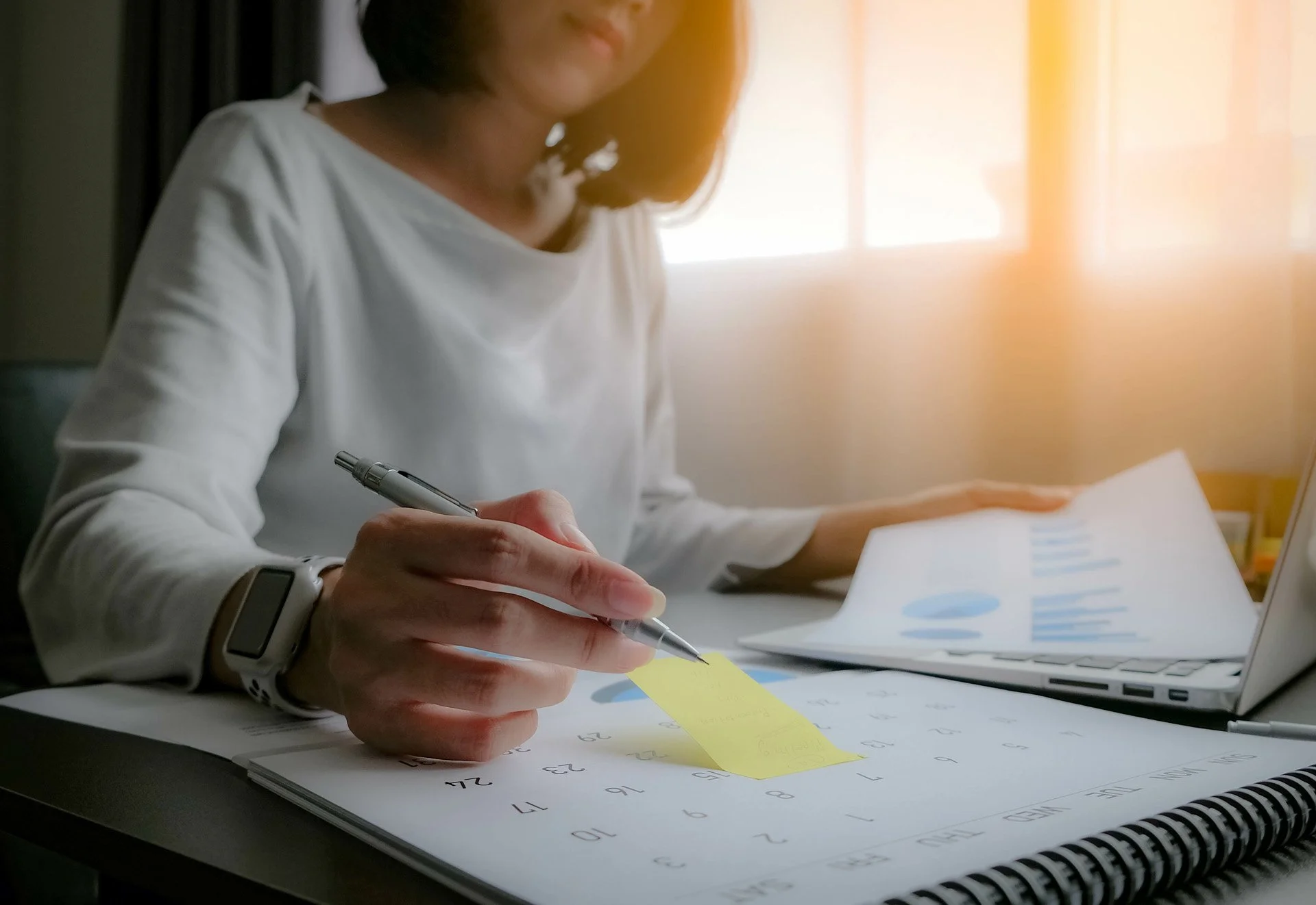 A woman sitting at a desk working with papers, a laptop, and a calendar; she holds a pen and a yellow sticky note.