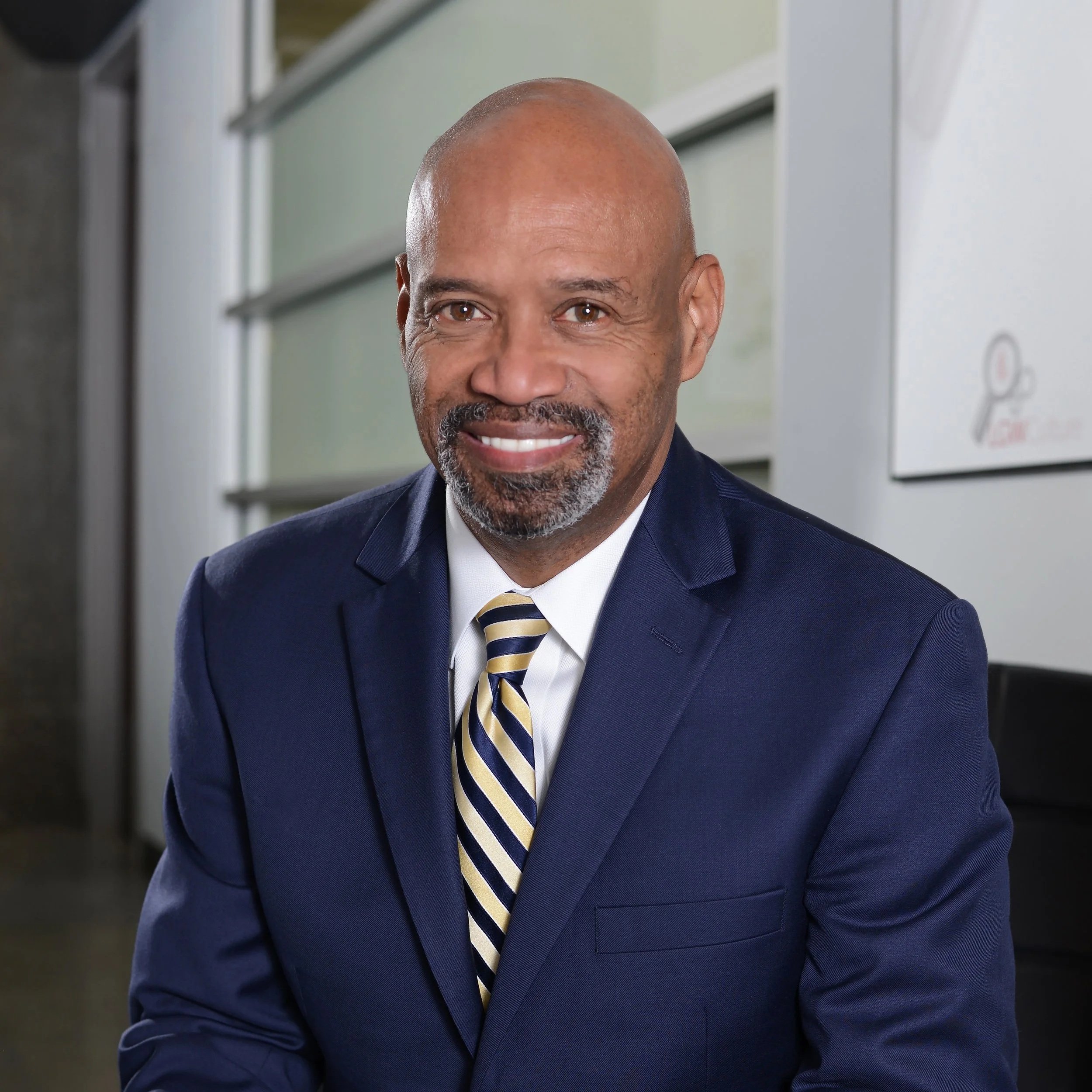 A smiling middle-aged African American man dressed in a navy suit with a striped tie, sitting in an office setting.