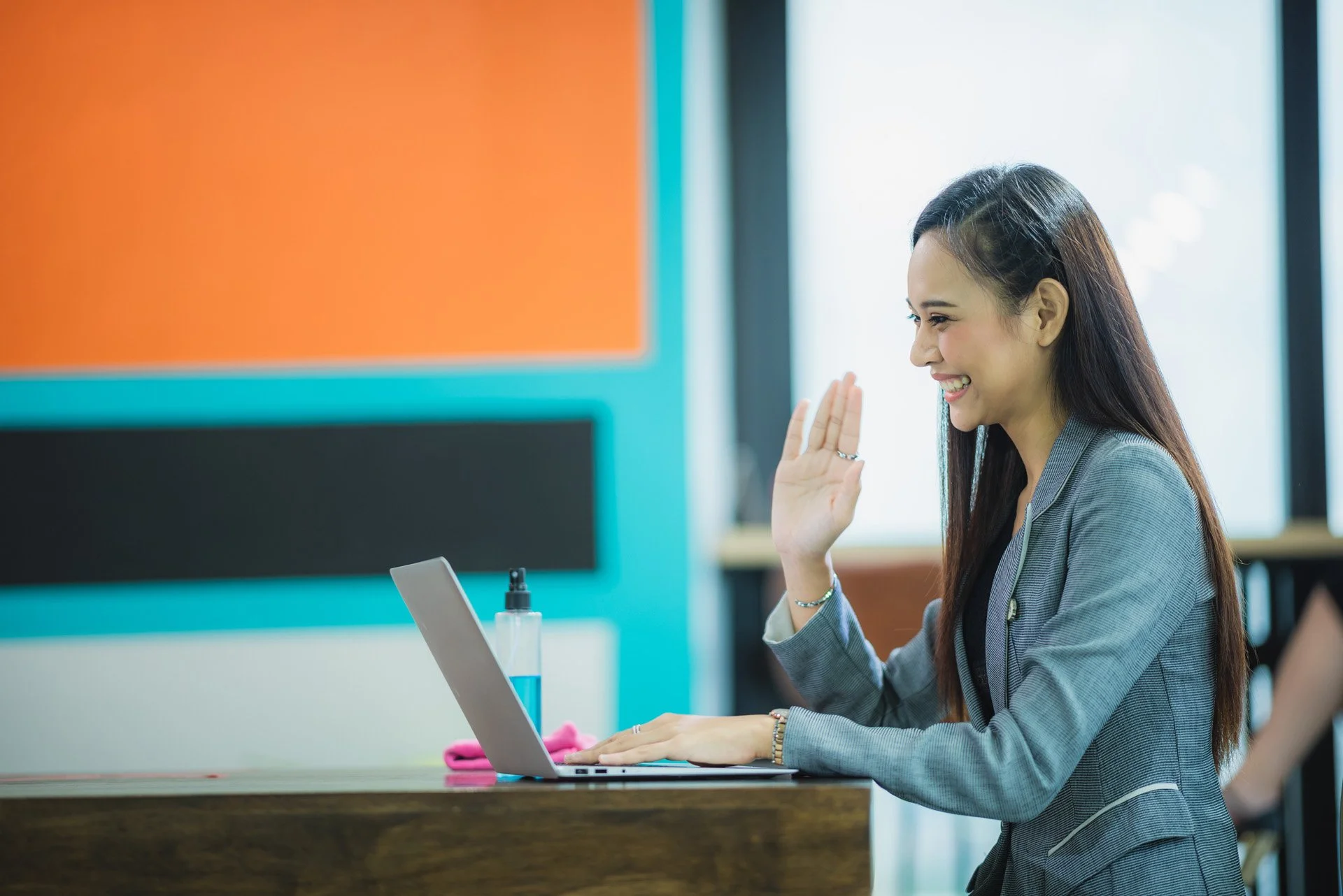 A woman in a gray blazer smiling and waving at her laptop in a modern office.