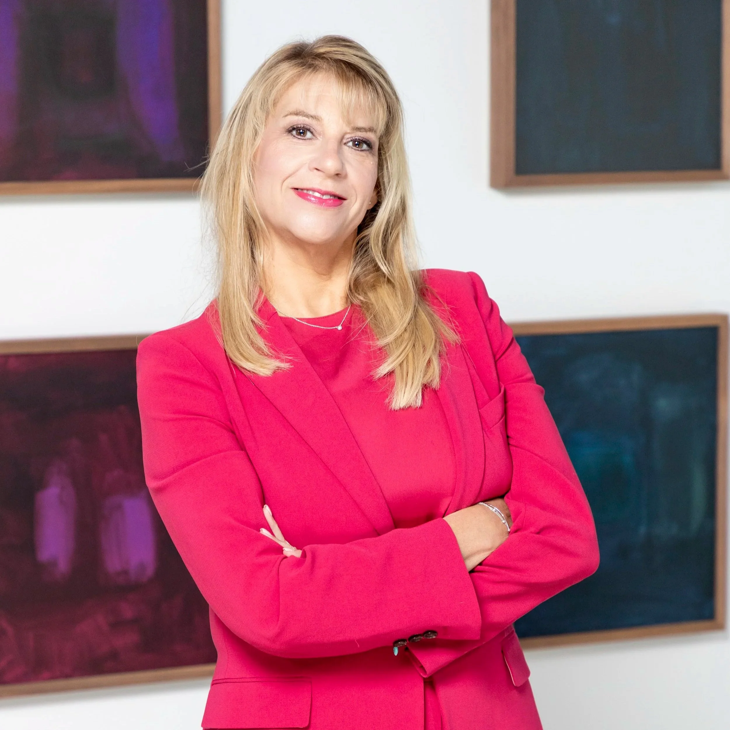 A confident woman with blonde hair wearing a red suit, standing with her arms crossed in front of abstract artwork on a white wall.