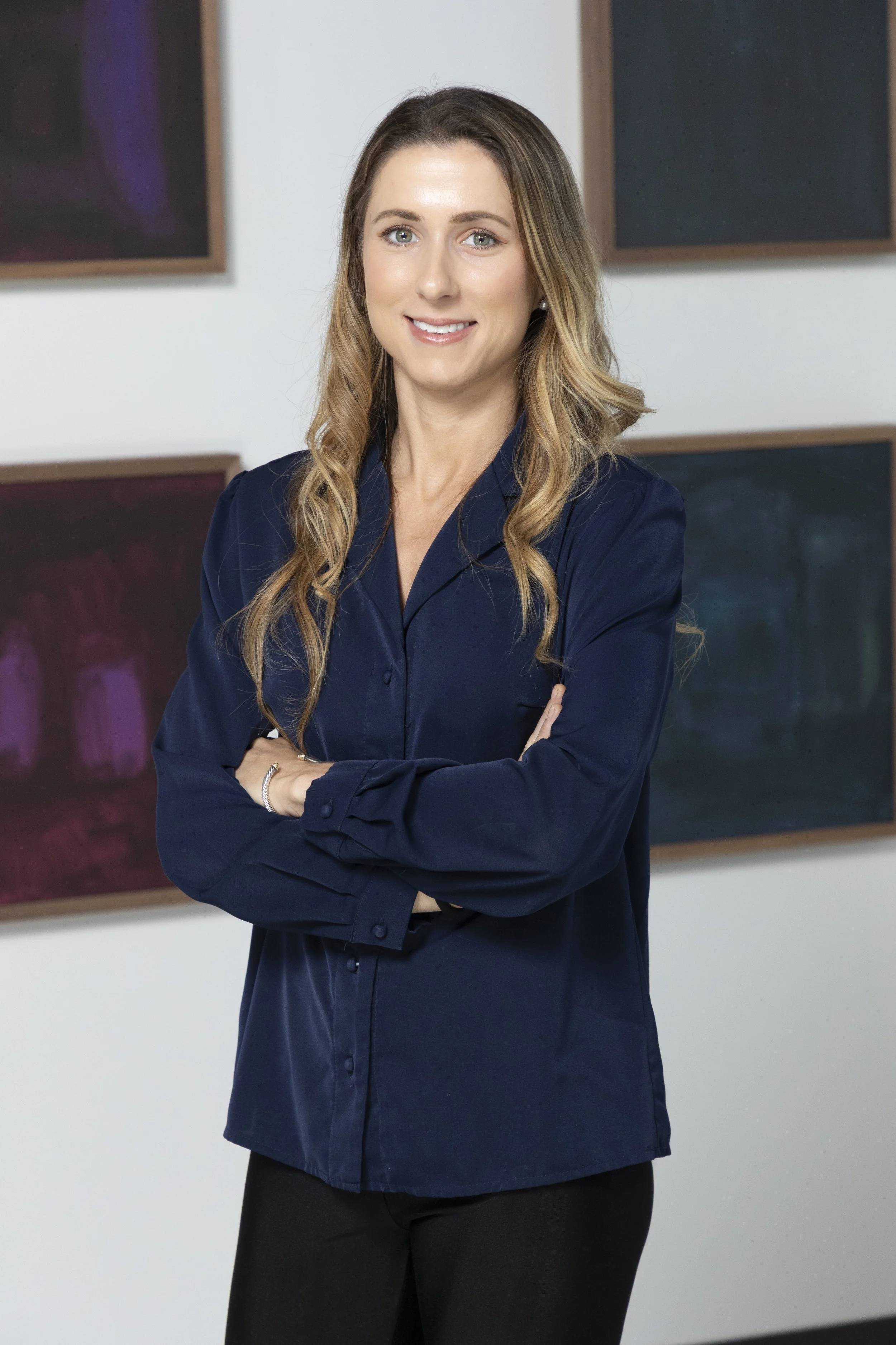 A woman with long light brown hair, wearing a dark blue button-up shirt and black pants, standing with arms crossed in front of a white wall with abstract artwork.