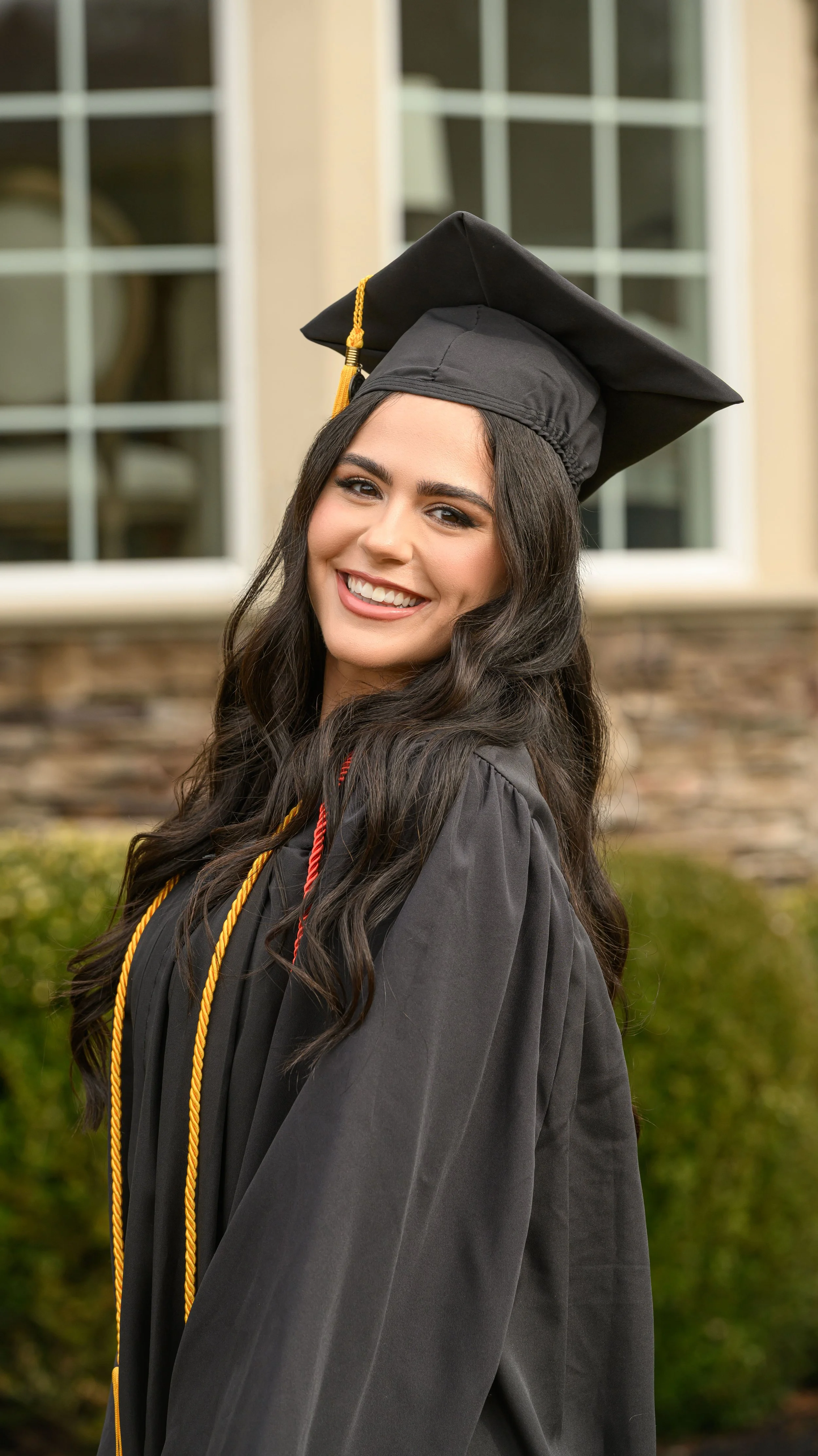 A young woman in a black graduation gown and cap with a gold tassel, smiling outdoors in front of a building with large windows and greenery.