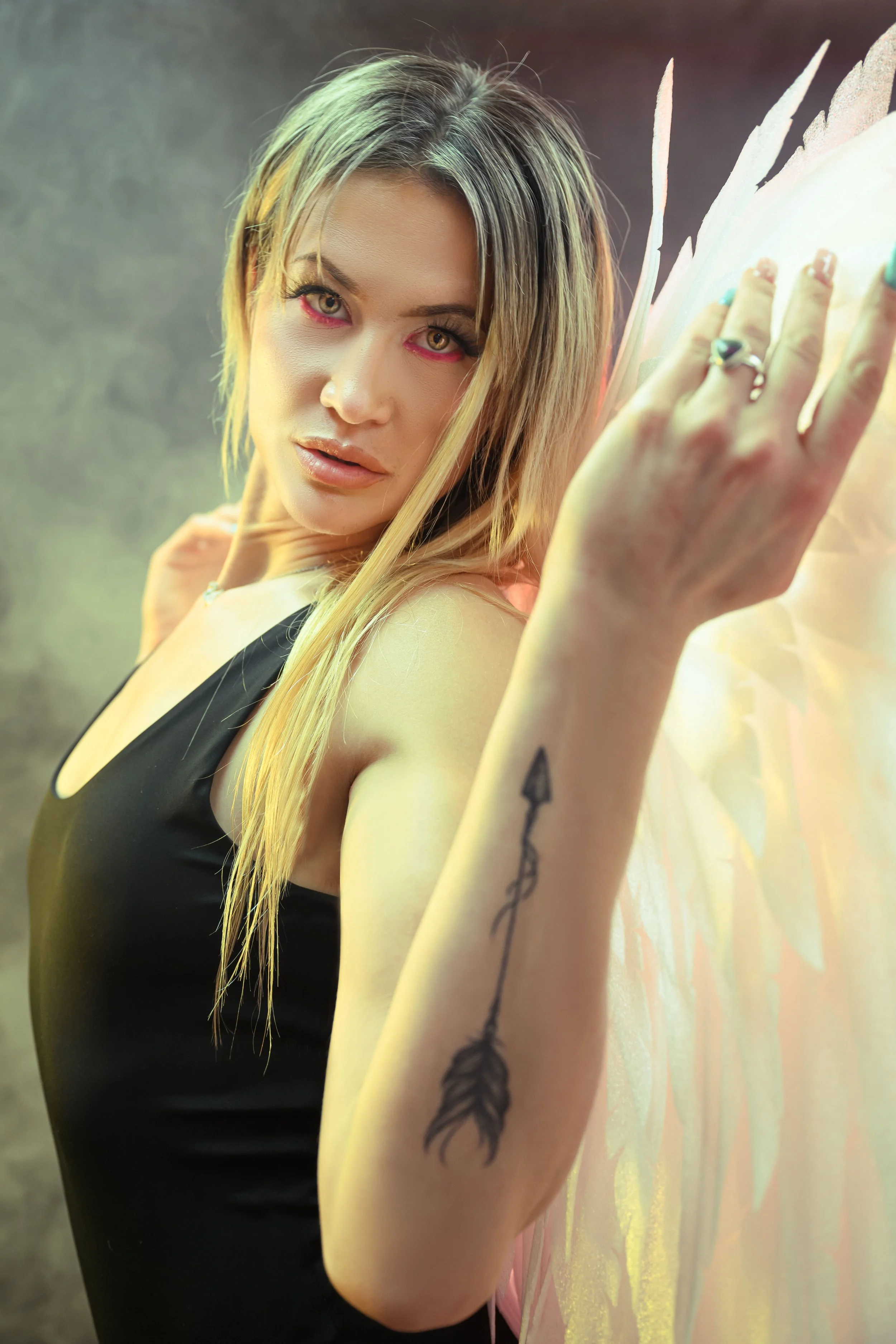 A woman with long blonde hair and fairy wings wearing a black tank top, showing a tattoo of an arrow on her forearm, looking at the camera with a slight smile.