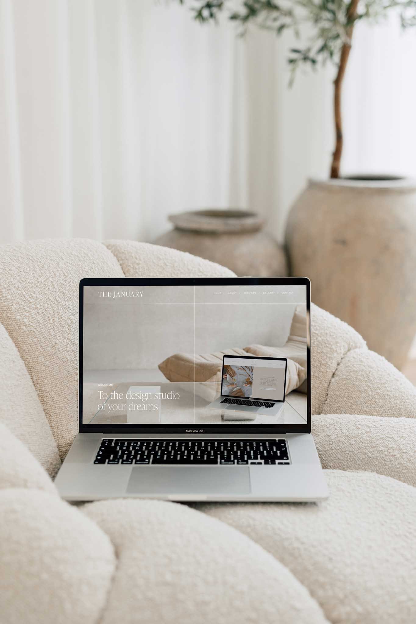 A MacBook Pro laptop on a cream-colored textured sofa, displaying a website titled 'The January' with the text 'To the design studio of your dreams' on the screen. In the background, there are two large beige ceramic pots and a potted tree with green leaves.