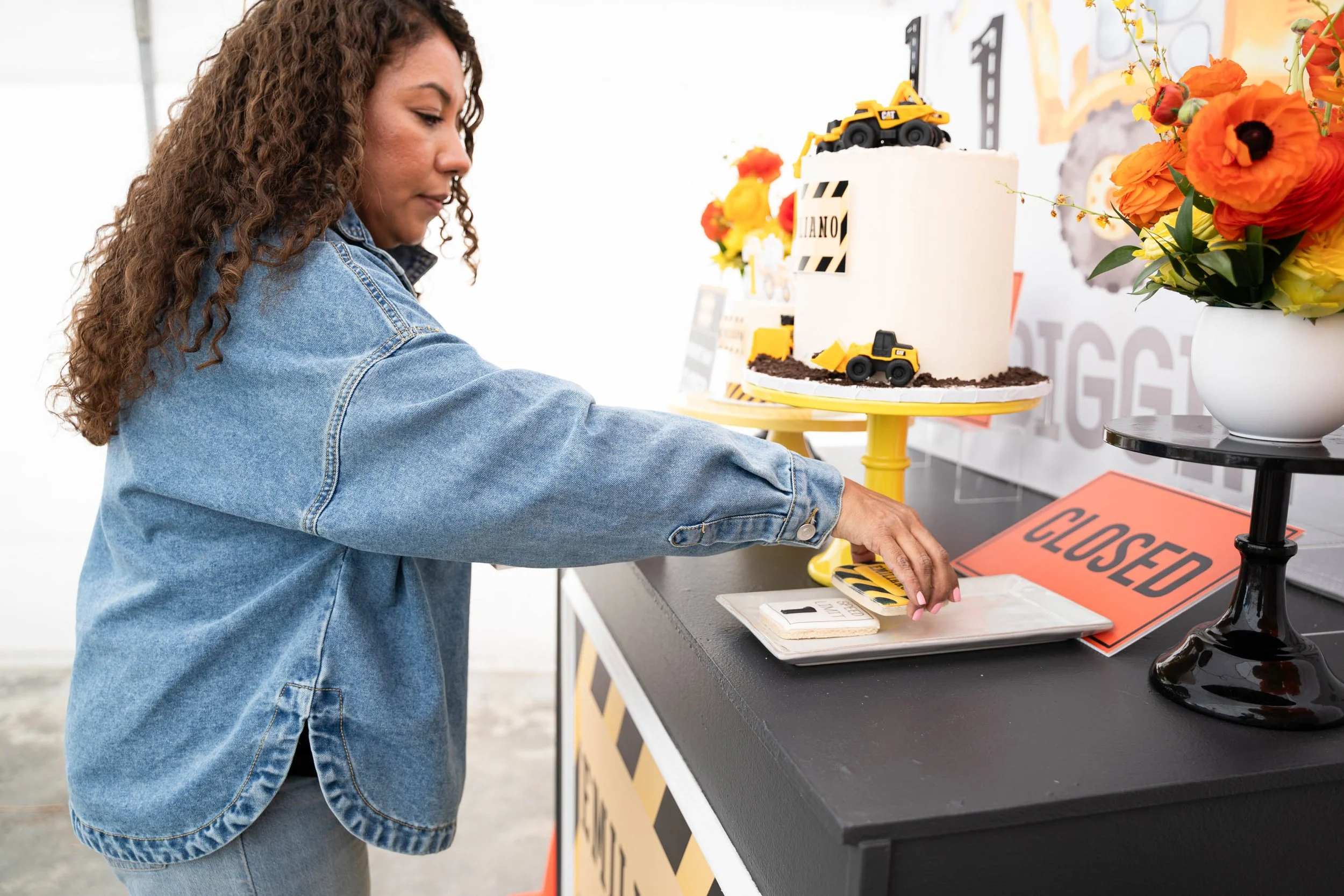Woman in denim jacket reaching for items on a table at a construction-themed birthday party decorated with a cake, construction toys, and a 'CLOSED' sign.