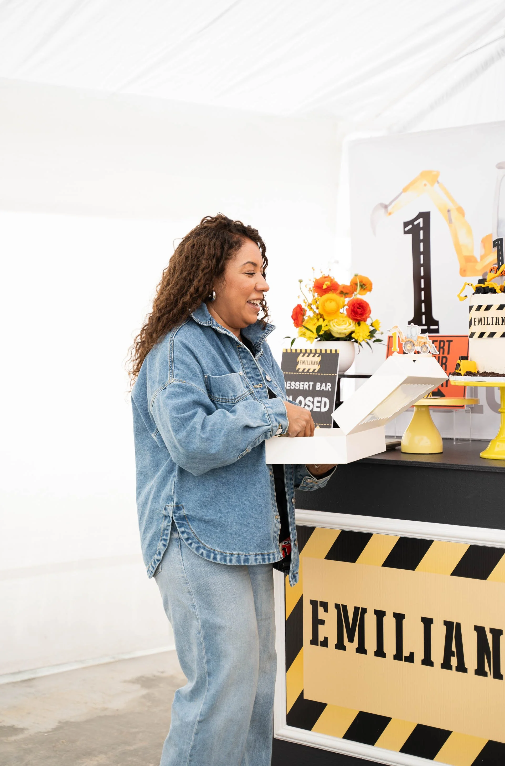 A woman in a denim jacket and jeans opening a cake box at a celebration table with flowers and birthday decorations.