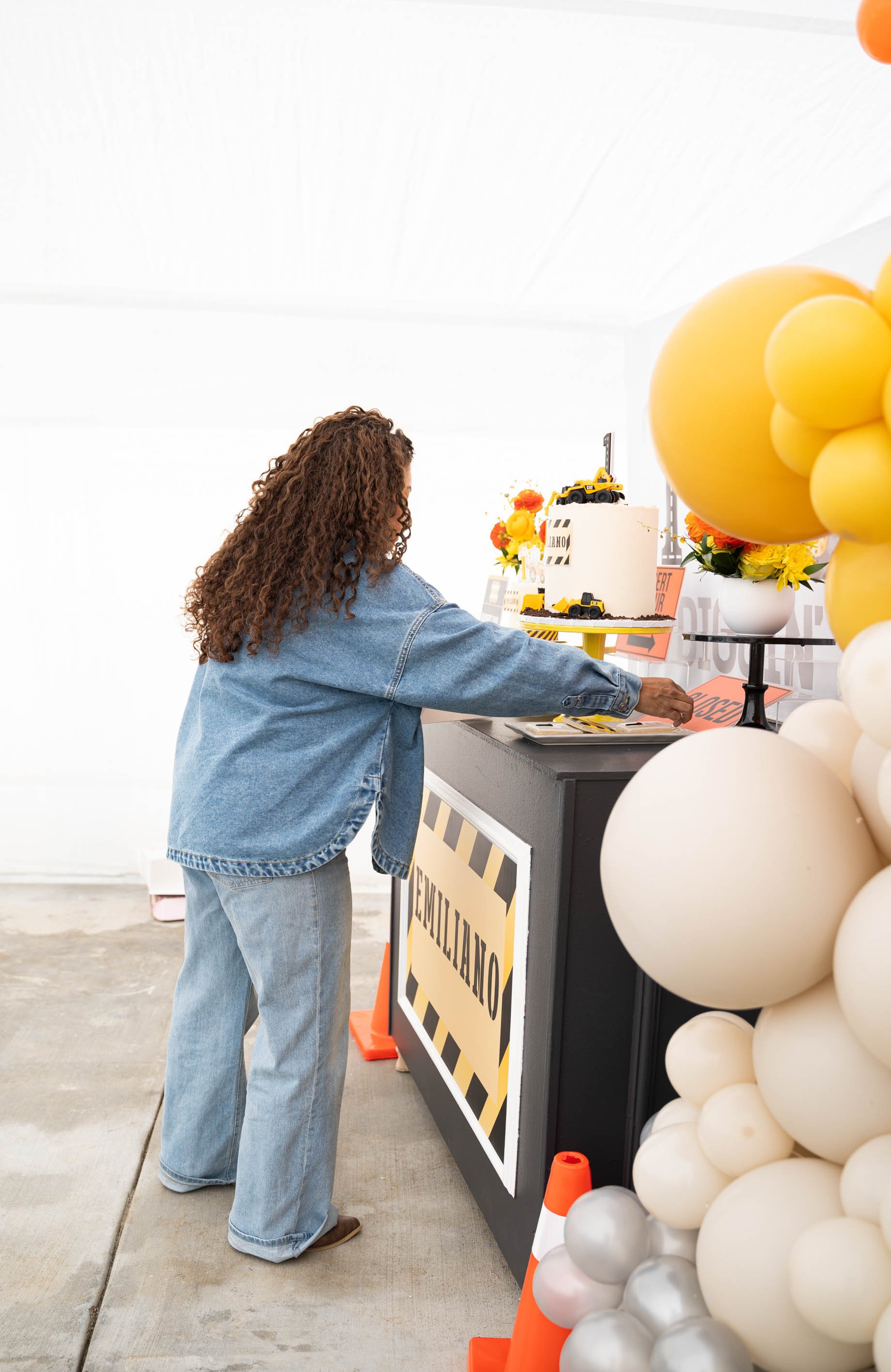 A woman with curly hair wearing a denim jacket and jeans is reaching for a cake at an emergency-themed celebration. The cake has construction vehicle decorations and is on a black table decorated with yellow and black caution tape signs. There are orange traffic cones and a balloon arrangement with yellow, white, and silver balloons nearby.
