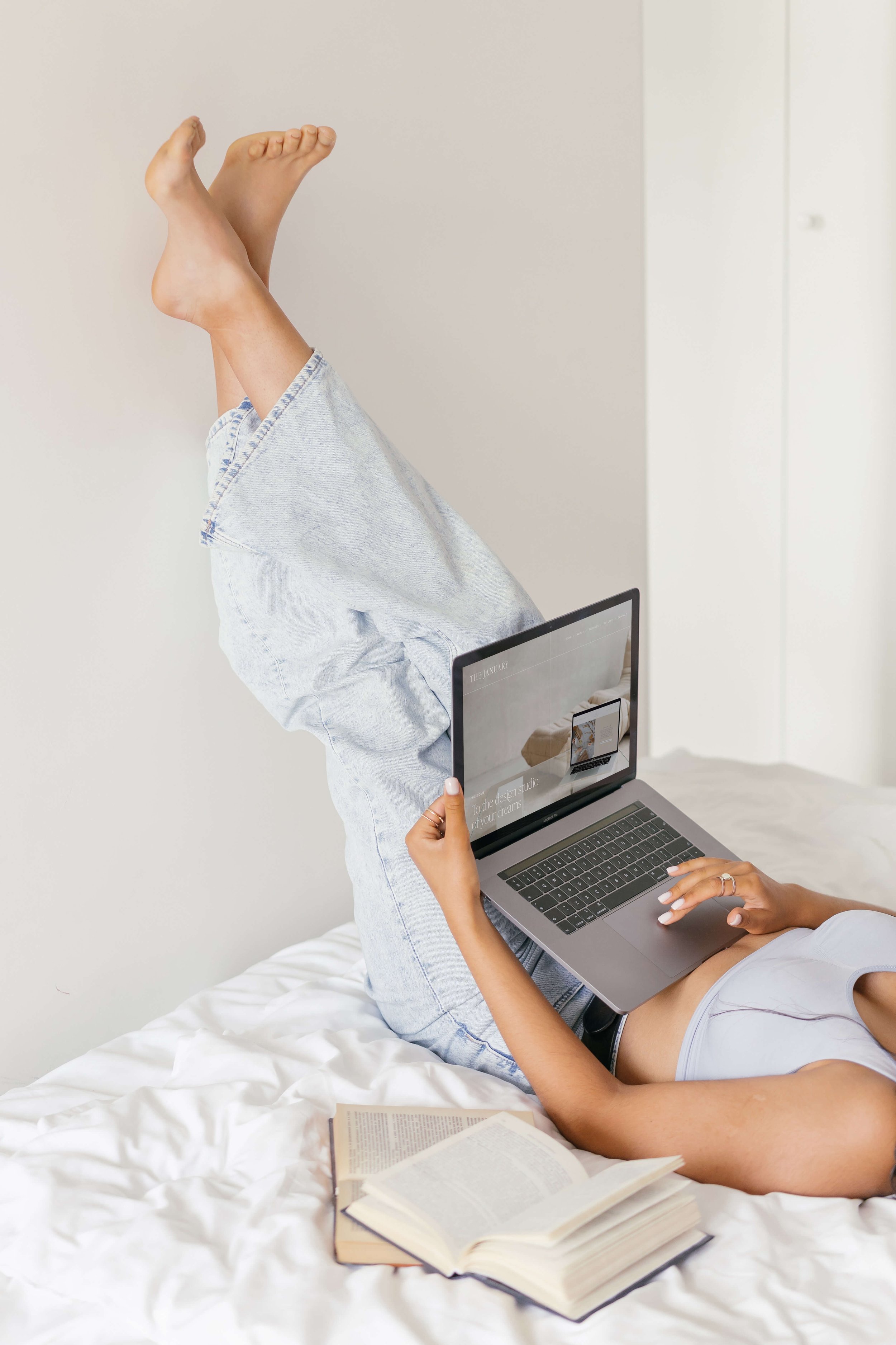 A person lying on a bed with legs up against a wall, holding a laptop and reading a book nearby.