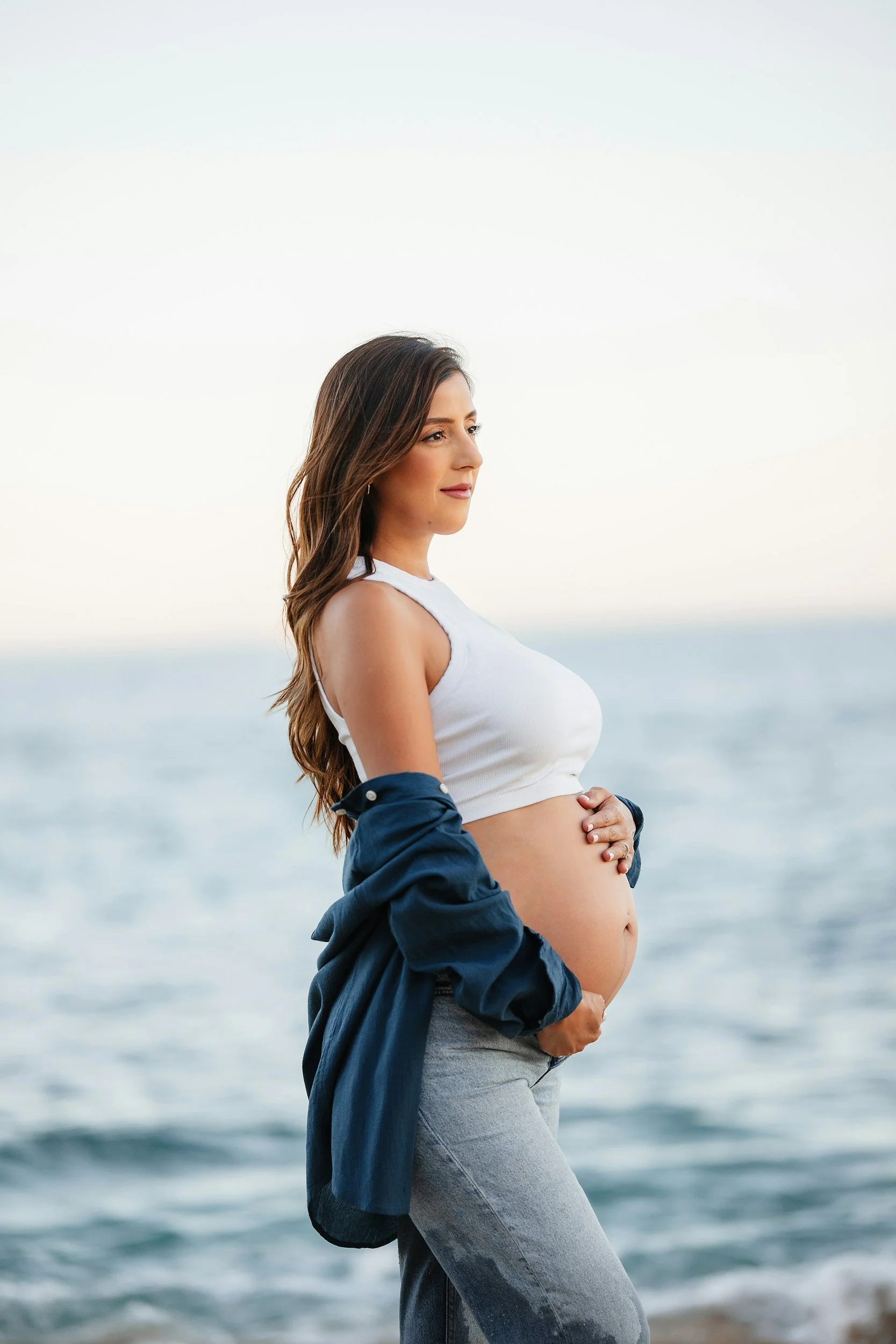 A pregnant woman standing at the beach with the ocean in the background, holding her belly with both hands, wearing a white sleeveless top and gray jeans.