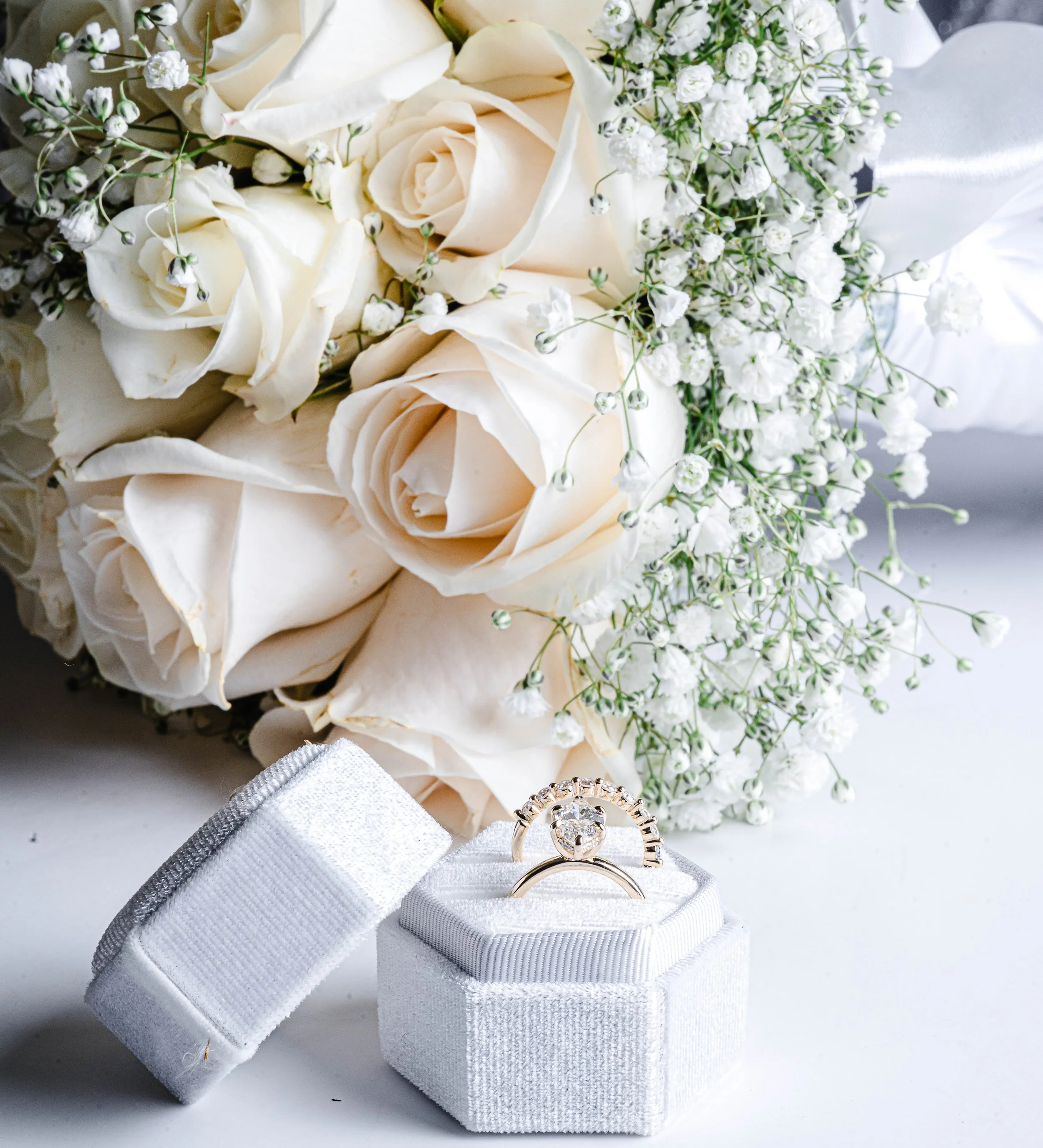 A bouquet of white roses and baby's breath flowers arranged together, with a diamond engagement ring and wedding band displayed on a white jewelry box next to it.