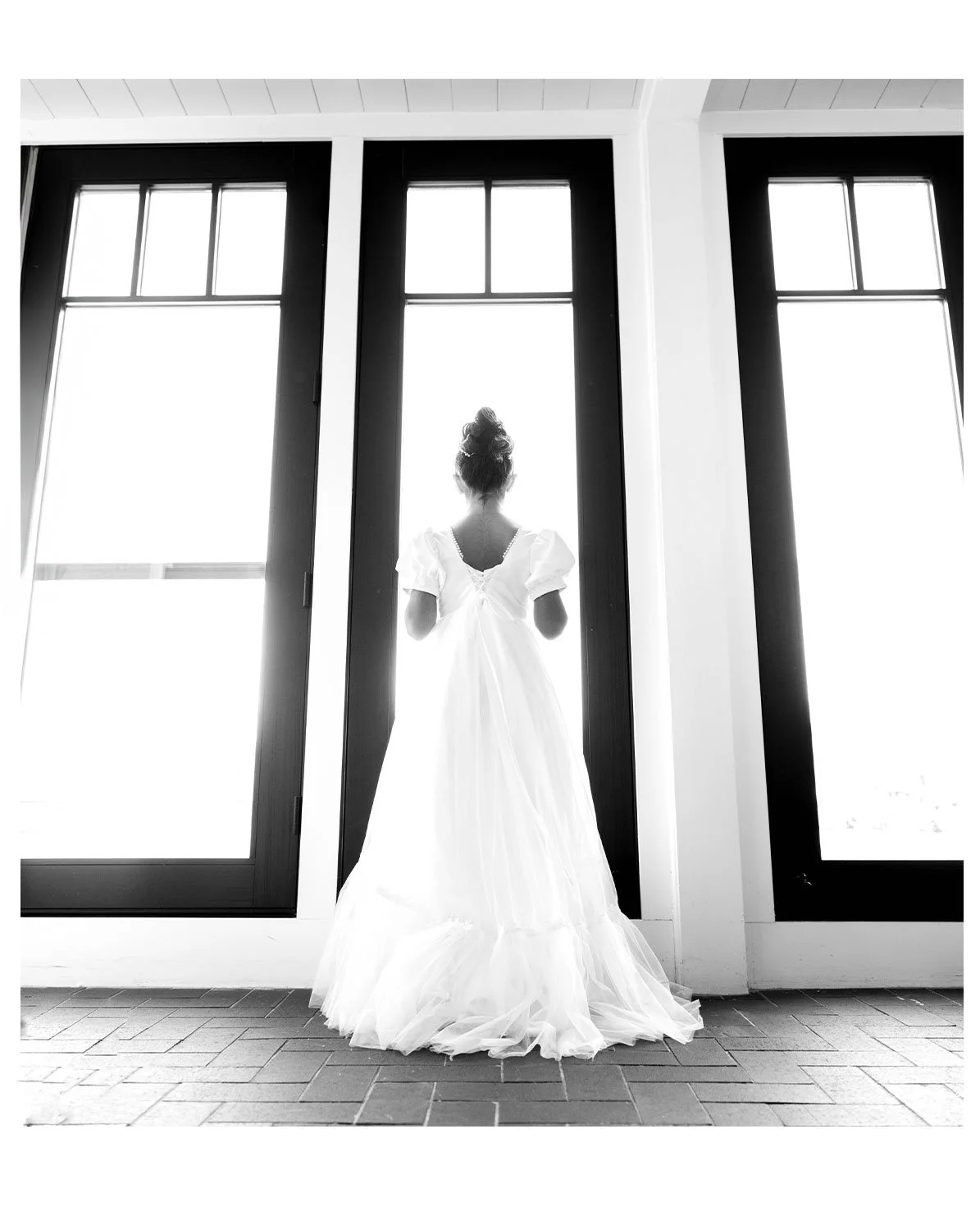 A woman in a white dress looking out large windows, back view, indoor setting.