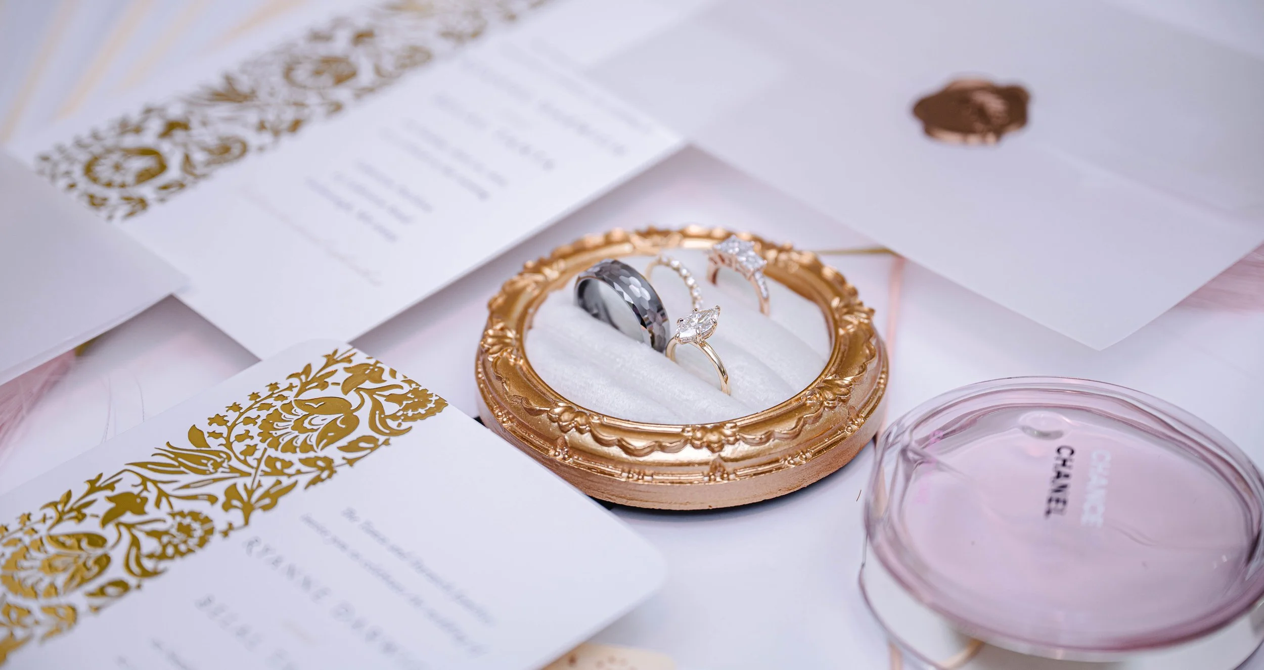 Close-up of a gold jewelry box with three rings inside, surrounded by wedding invitations and a pink Chanel makeup compact.