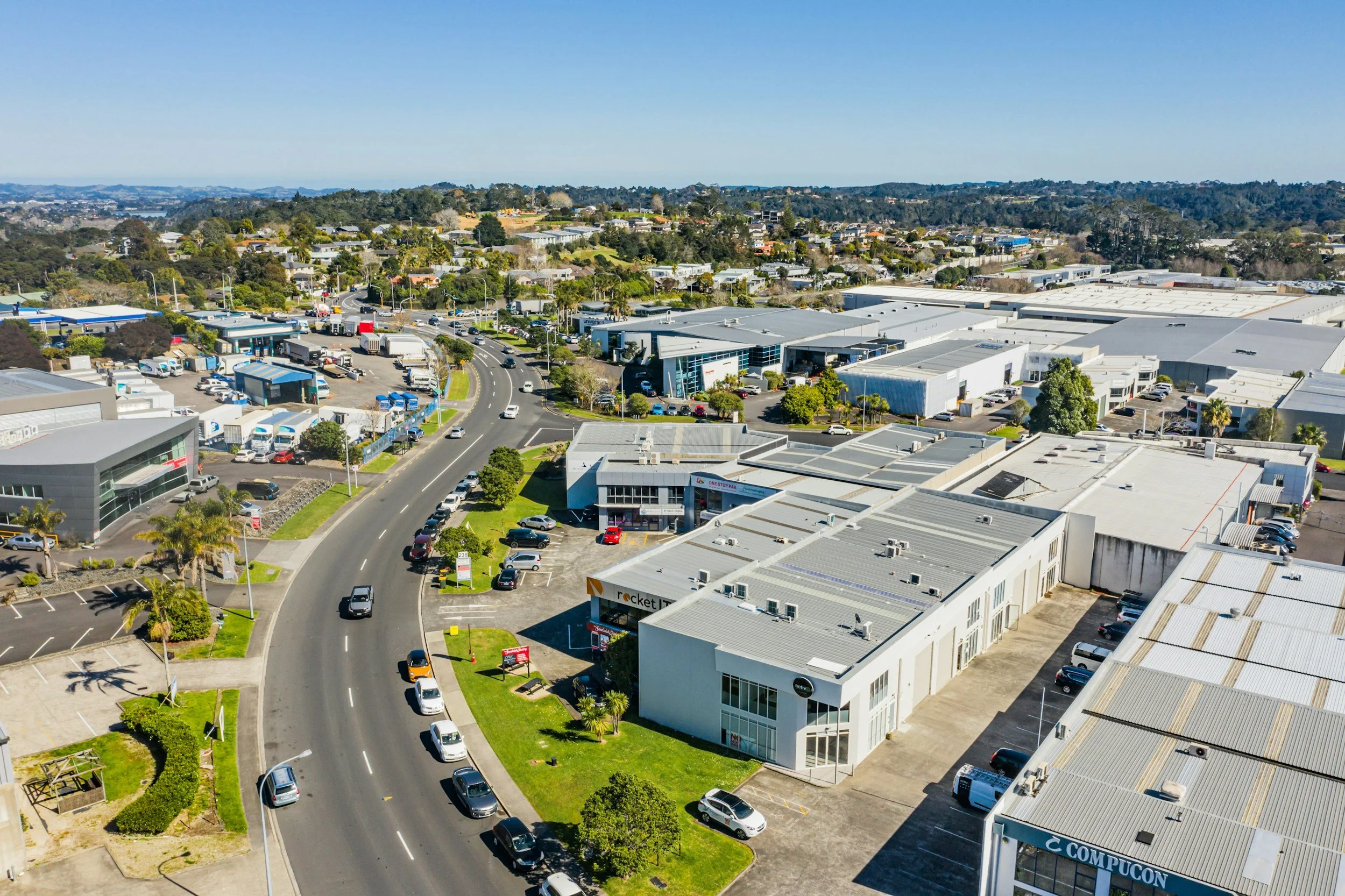 Aerial view of a commercial area with roads, shops, and parking lots, surrounded by greenery and distant hills under a clear blue sky.