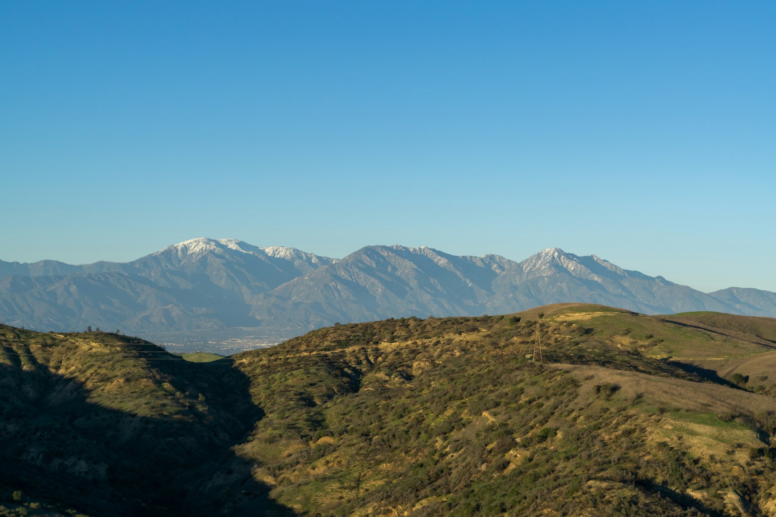 Scenic view of rolling green hills with patches of grass and shrubs, mountain range with snow-capped peaks in the background, under a clear blue sky.