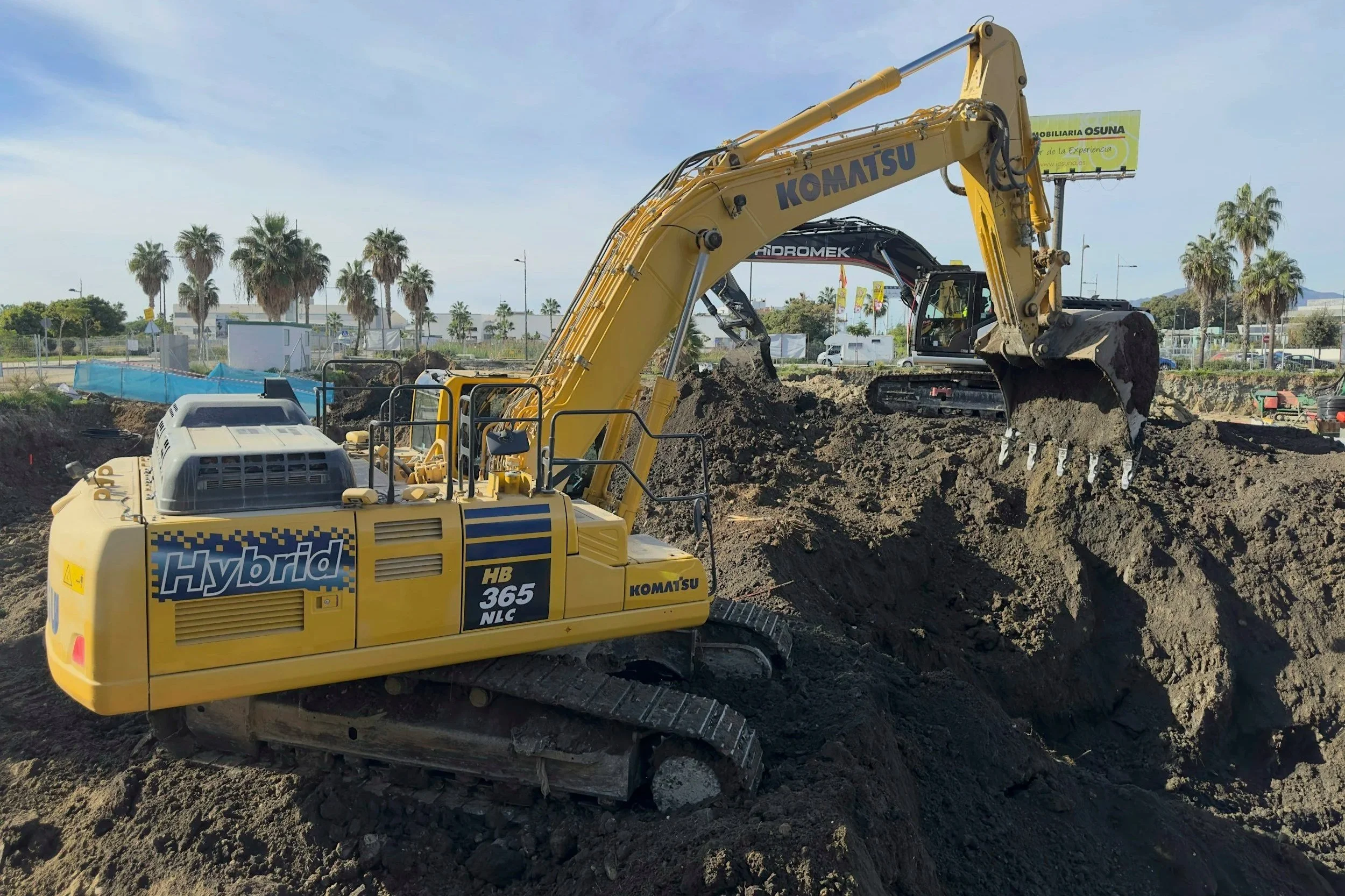 Yellow Komatsu hybrid excavator digging in a construction site with palm trees and a billboard in the background.