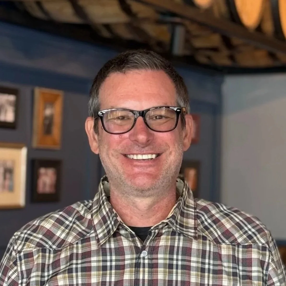 A smiling man with glasses wearing a plaid shirt, standing indoors with framed pictures on the wall behind him.